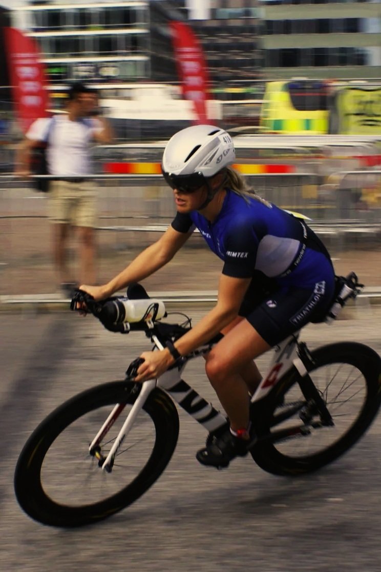 Psykolog och triathlet tävlar i triathlon. Hennes hår fladdrar i vinden, hon bär hjälm och sportkläder, med bakgrund av stadsmiljö och personer.