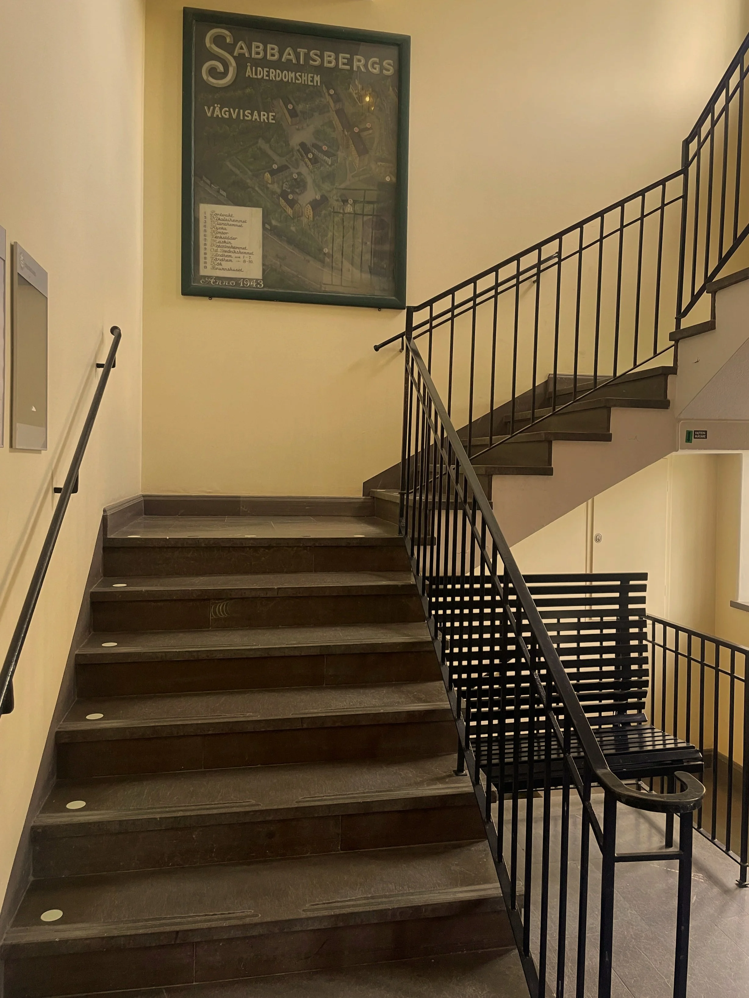 En trappa med trappsteg av mörkt trä och svart metallräcke, en vägg med en grön ram på en palst och en liten dörr nedanför trappan.