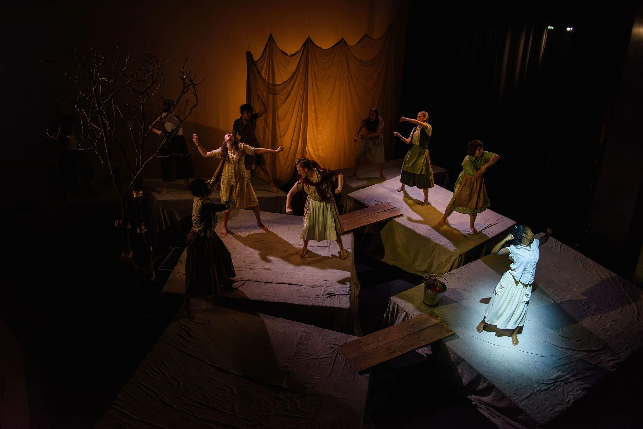 Theater scene with actors on stage, some standing, some sitting, with a backdrop and some props, illuminated with warm stage lighting.