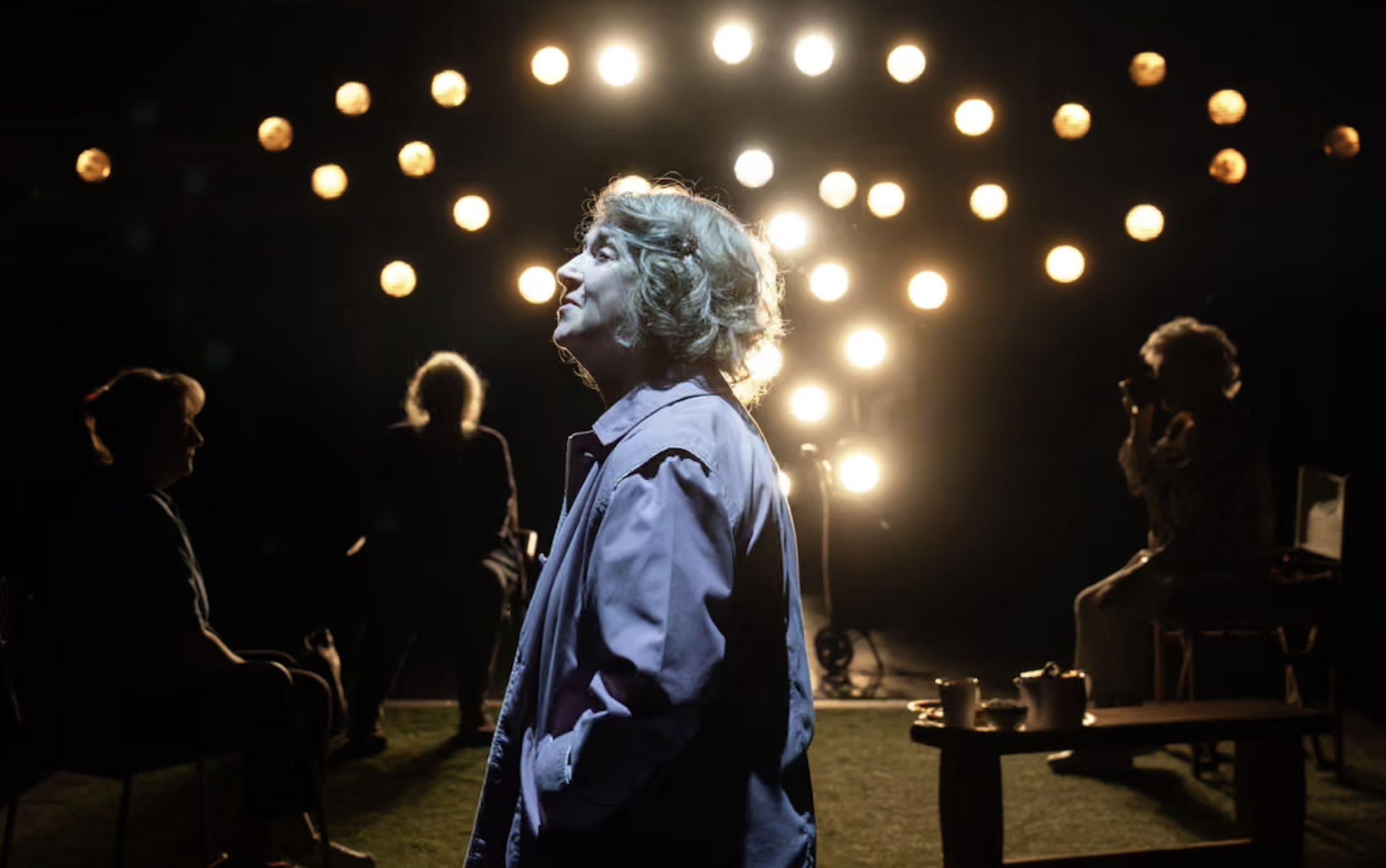 Woman with curly hair standing in the spotlight during a performance or rehearsal, with other figures in the background, illuminated by bright stage lights.