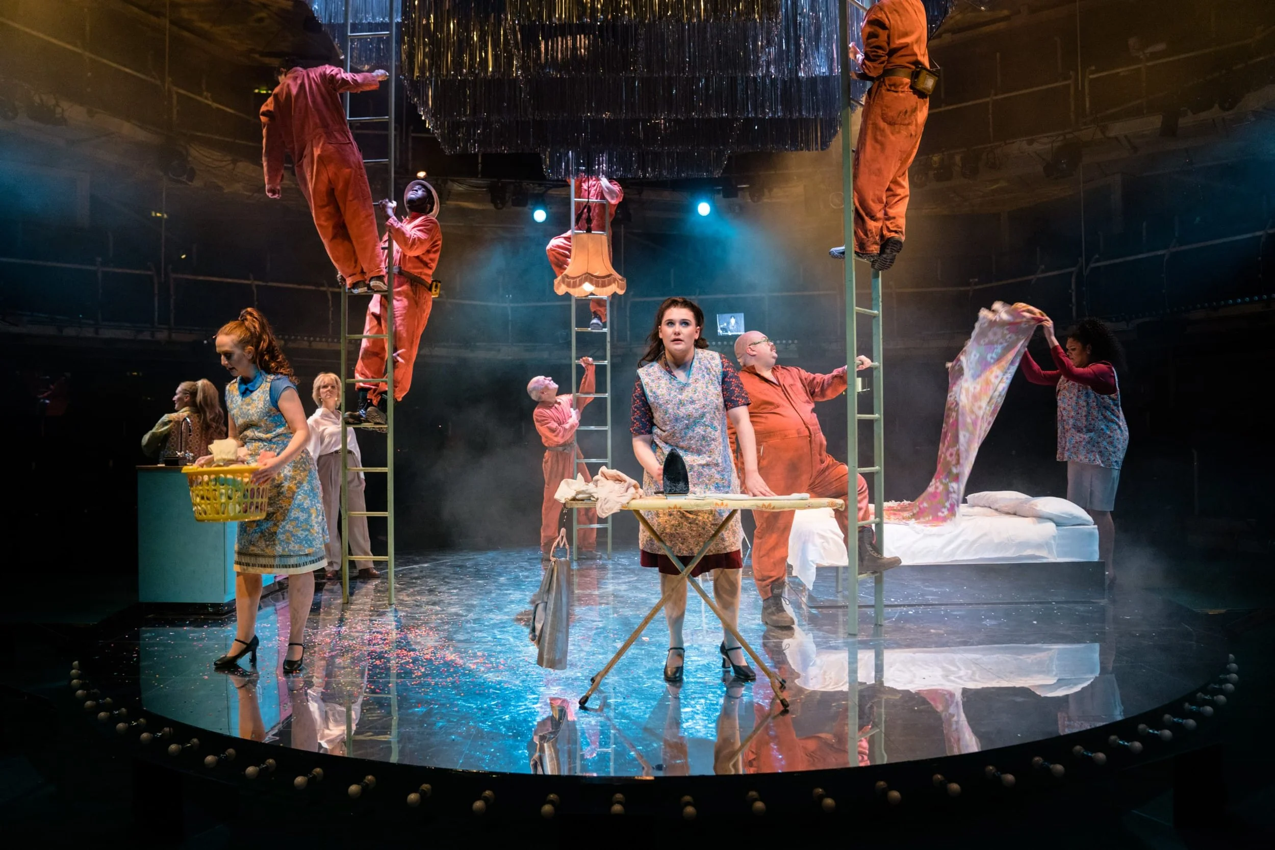 The image shows a theatrical stage with actors in costume performing a scene. Some actors are on ladders hanging upside down, others are standing or sitting, engaging in various activities like ironing, folding fabric, and arranging items. The stage is decorated with a large chandelier hanging overhead, and there is a reflective floor with scattered confetti.