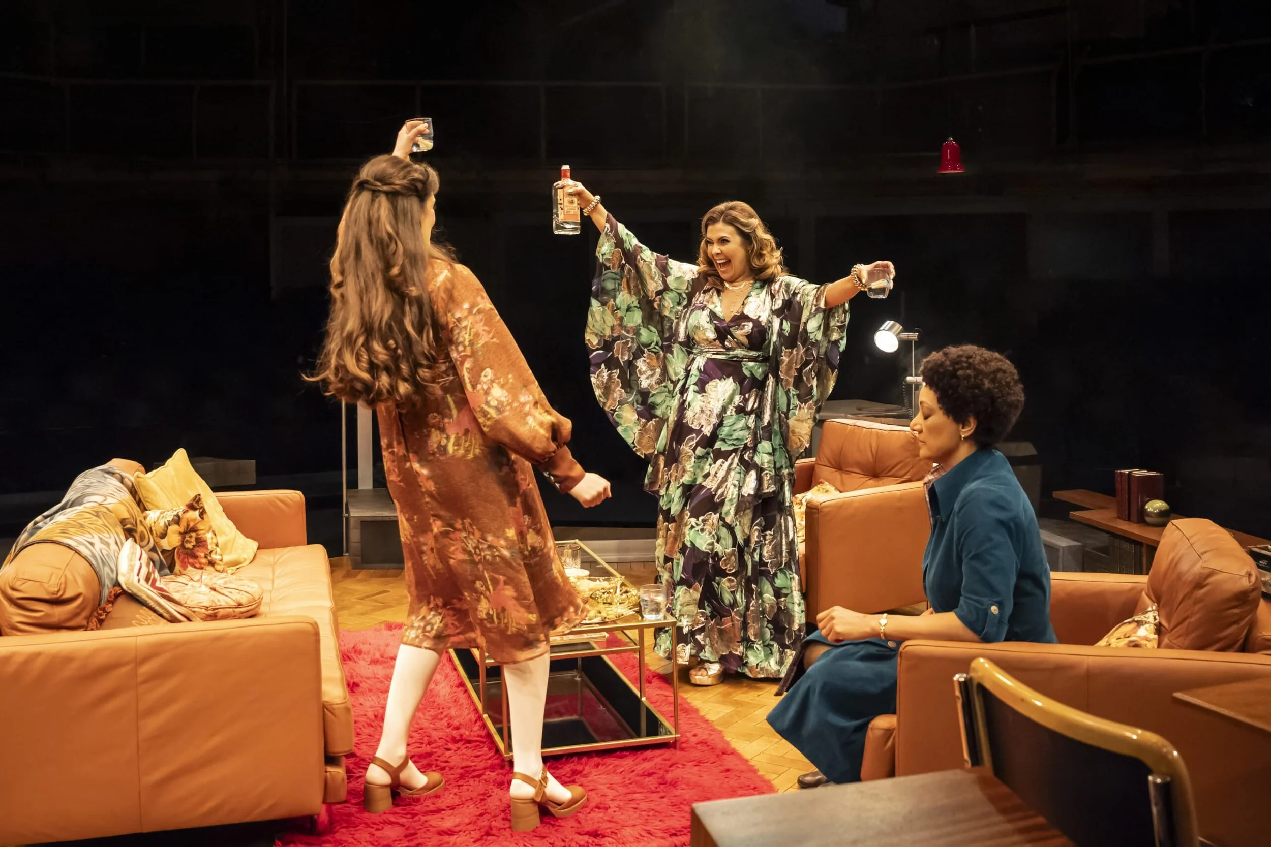 Four women celebrating in a cozy living room, with drinks raised, one standing and two seated on leather sofas, a coffee table with snacks and glasses, and a red rug on wooden floor.