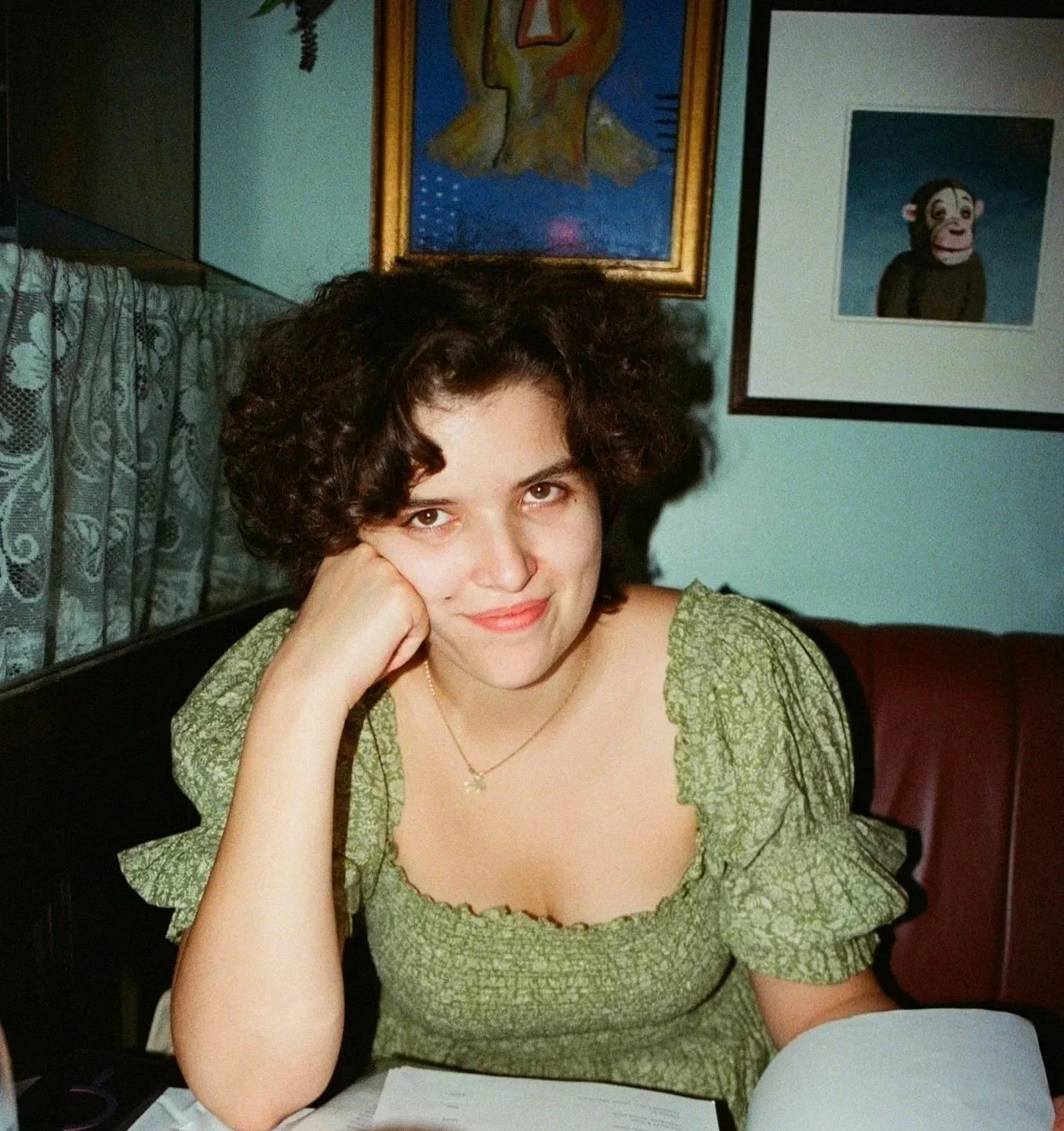 A woman sitting at a table with her head resting on her hand, smiling at the camera. She has curly dark hair and is wearing a green patterned dress and a necklace. Behind her are framed paintings on a light blue wall.