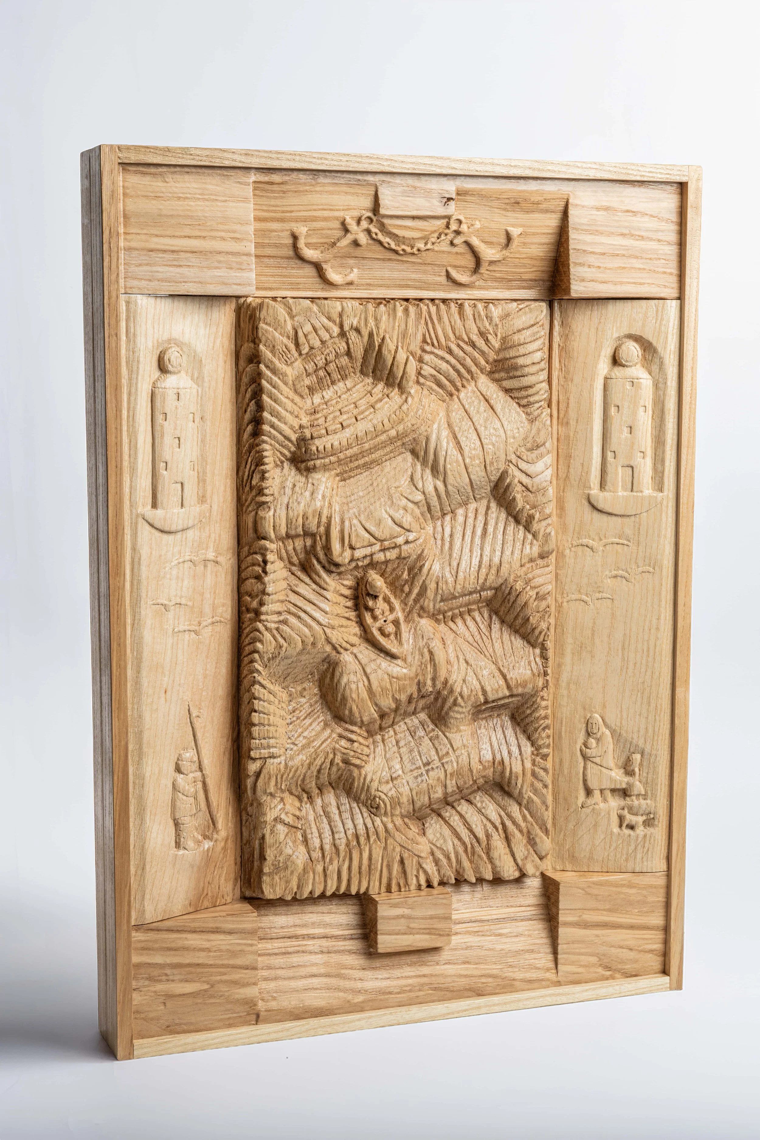 Wooden carved artwork depicting a figure with animal features, flanked by two lighthouse carvings, surrounded by a wooden frame with decorative elements at the top.