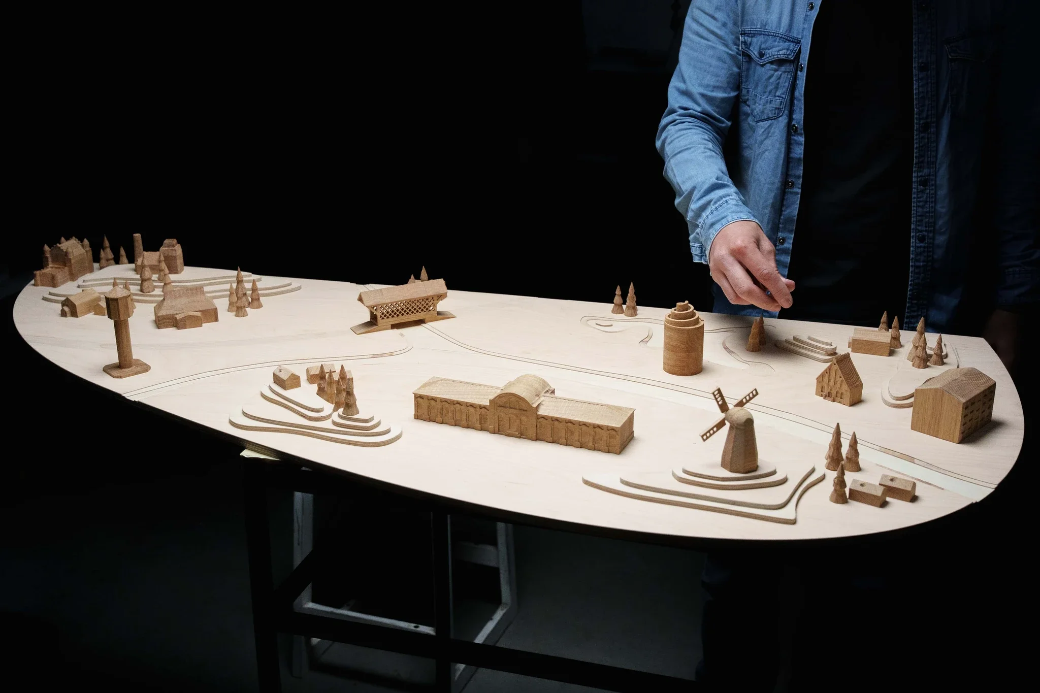 A person in a blue jacket is working on a detailed wooden model of a landscape with buildings, trees, a windmill, and pathways on a large table.