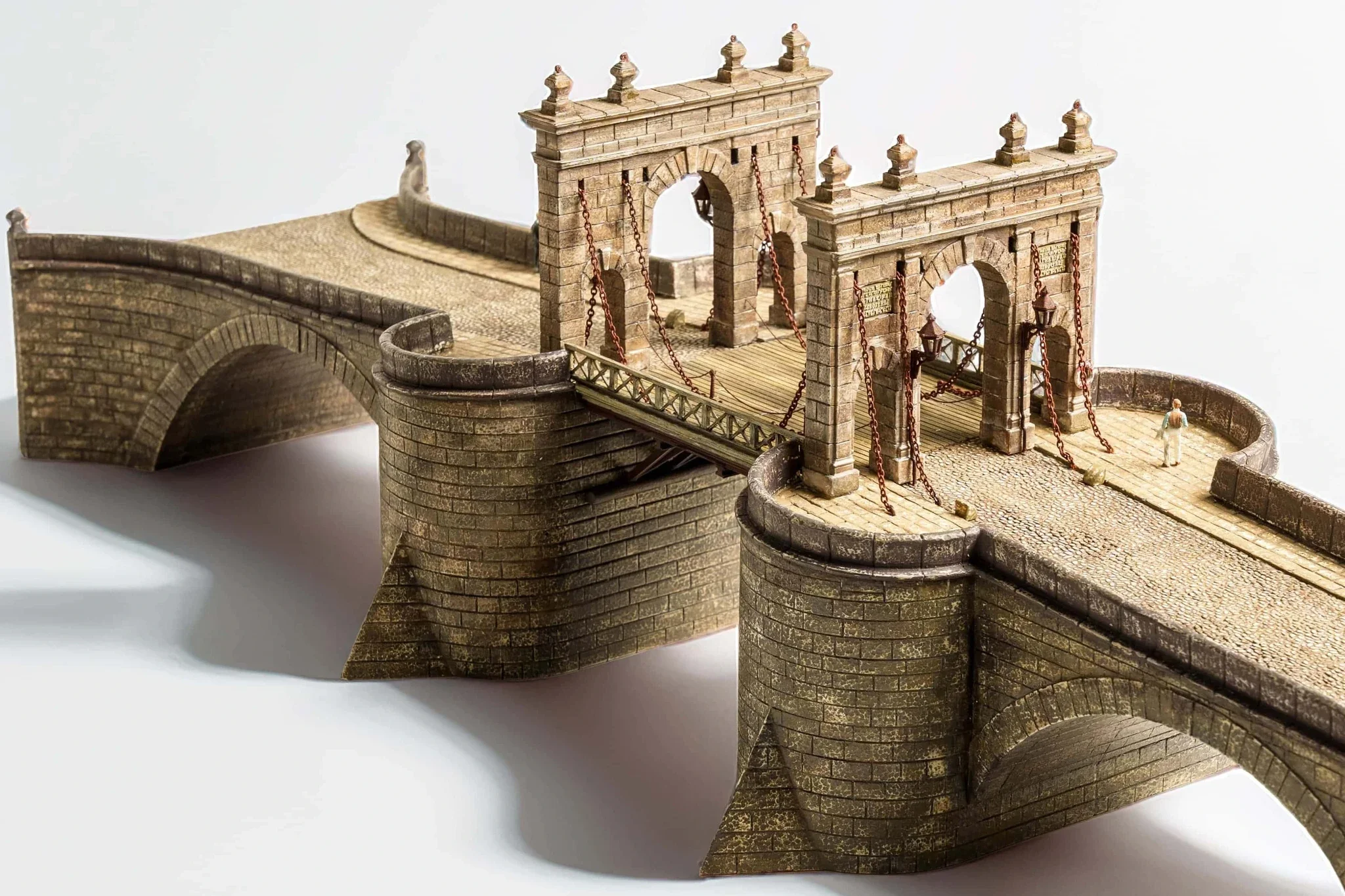 A detailed miniature model of a historic stone bridge with towers, arches, chains, and a wooden walkway. Small human figures are included for scale.