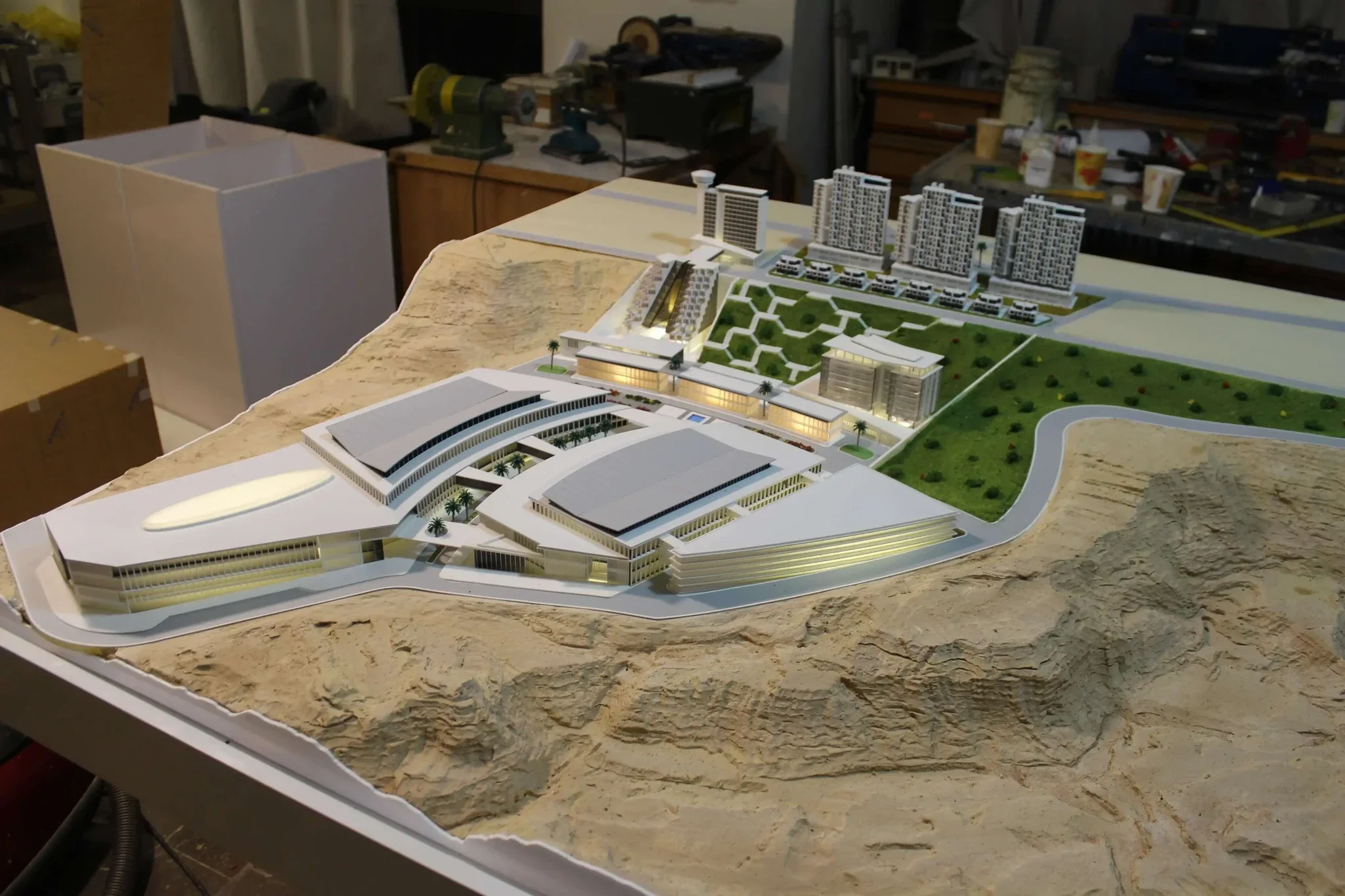 An architectural model of a large development project with multiple modern buildings, green spaces, and a road on a terrain with hills, displayed on a table in a workshop.
