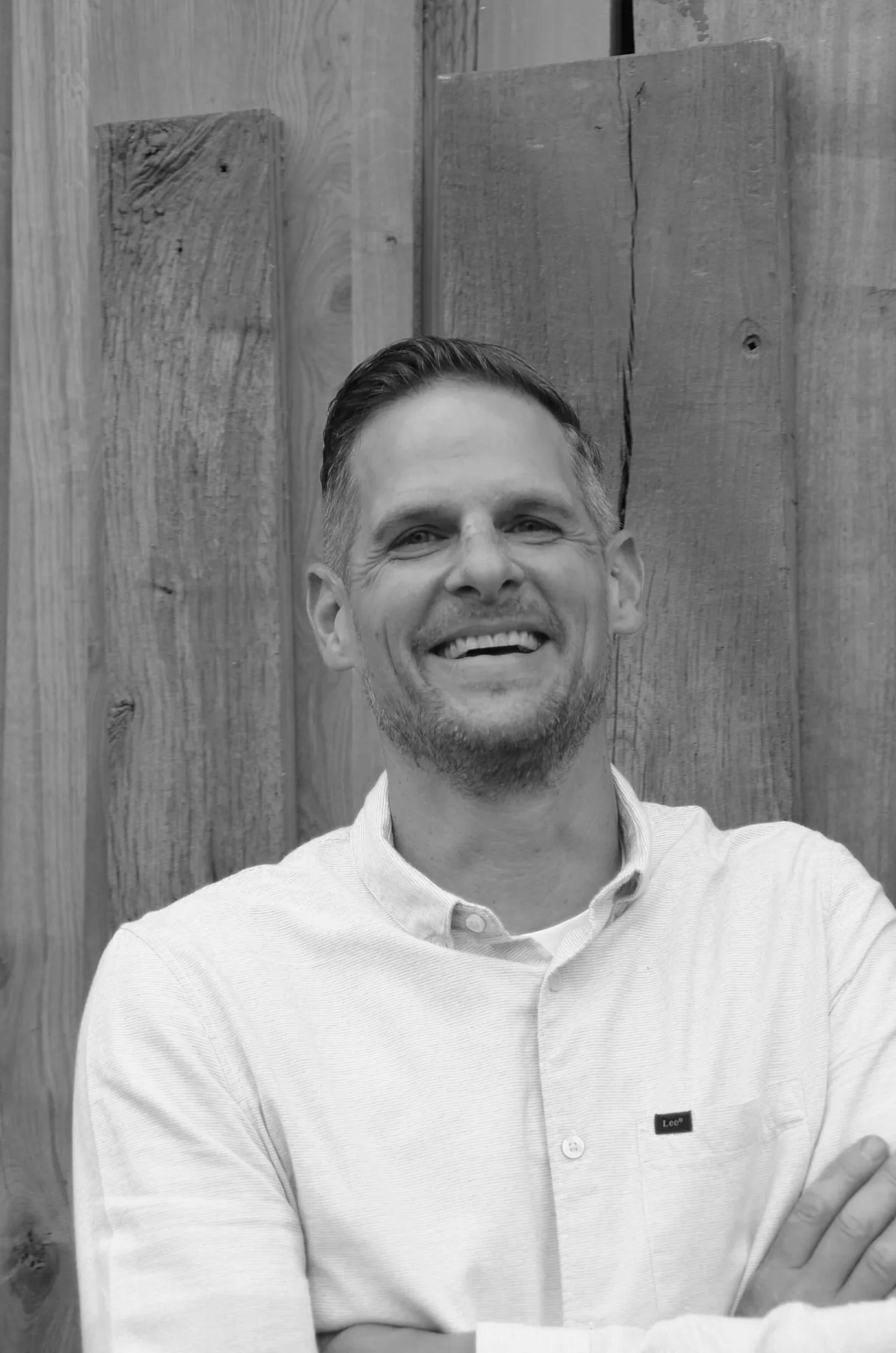 Black and white photo of a smiling man in a light-colored collared shirt standing in front of a wooden wall.