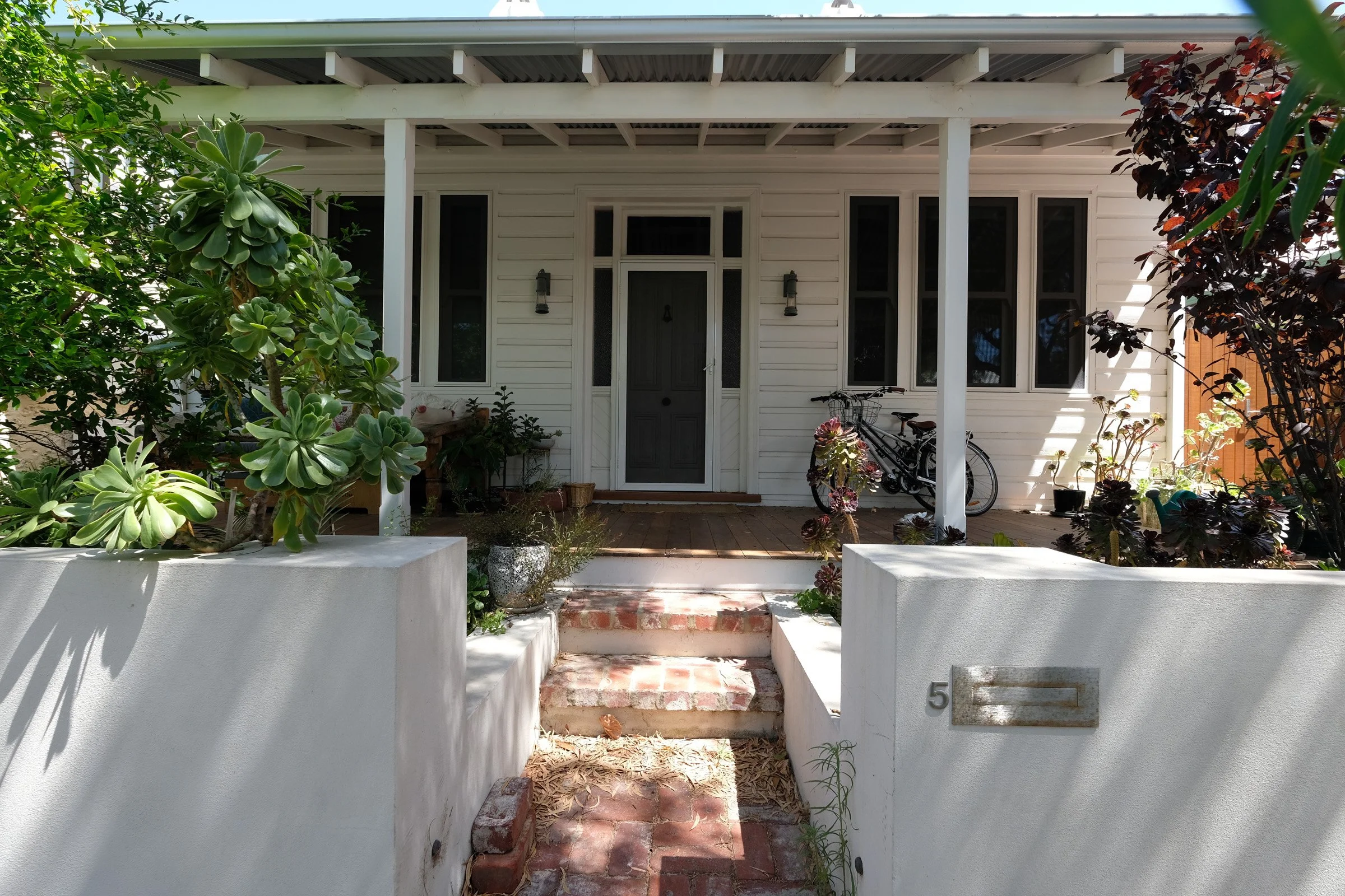 Front porch of a house with white siding, a central door, and bicycles on the right side. Plants and bushes surround the entrance, with brick steps leading up to the porch.