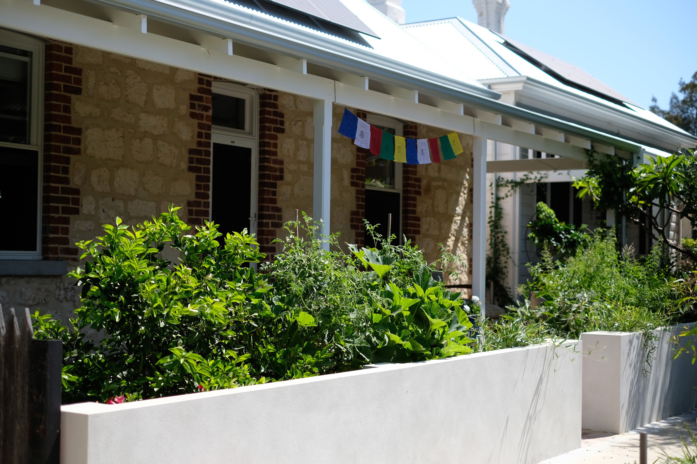 Outdoor balcony with seating area, white patio furniture, large white umbrella, wooden fence, greenery, and neighboring rooftops under a clear blue sky.