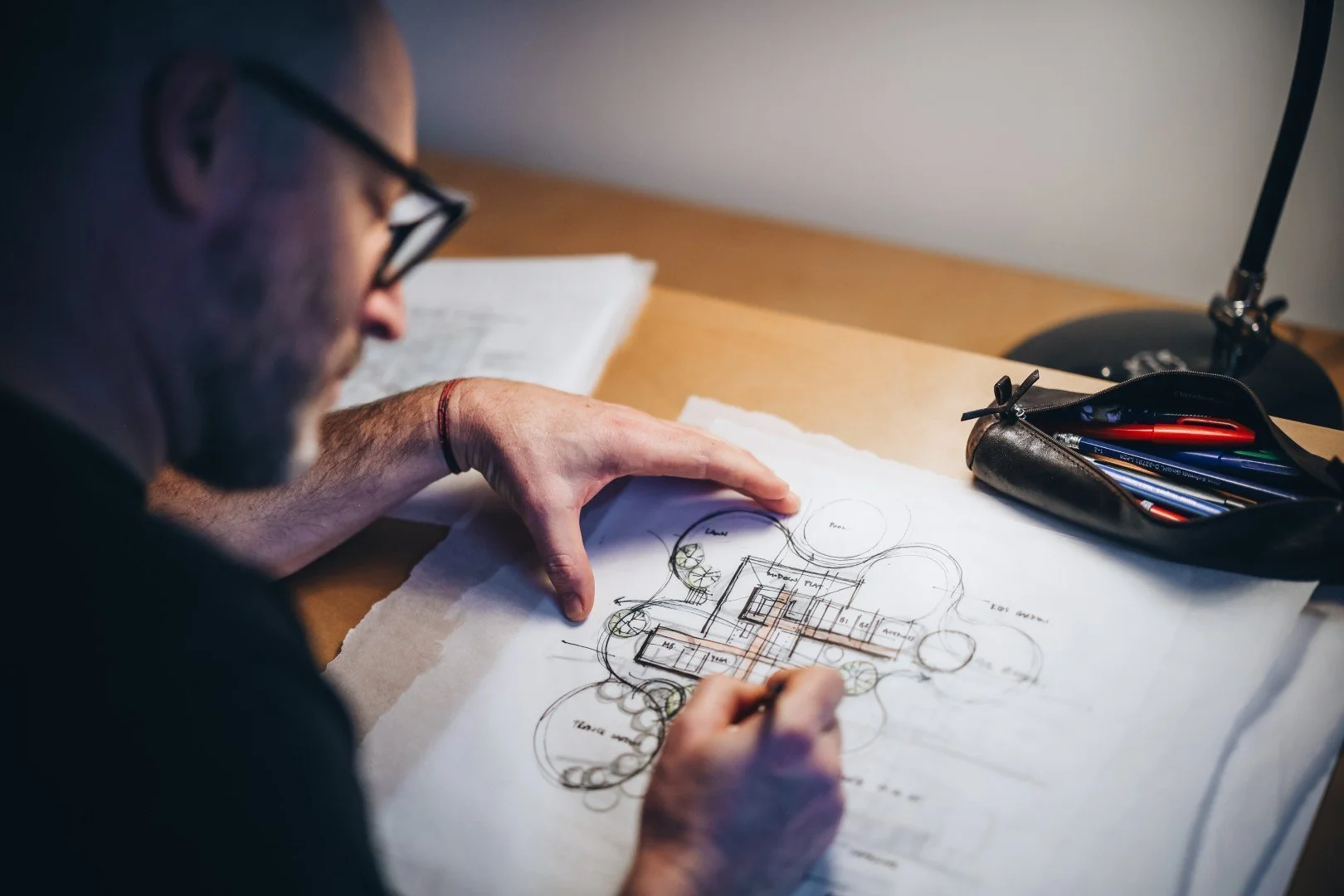 A man with glasses sketches a building layout on architectural plans at a desk, with a pencil in hand and a pen case nearby.