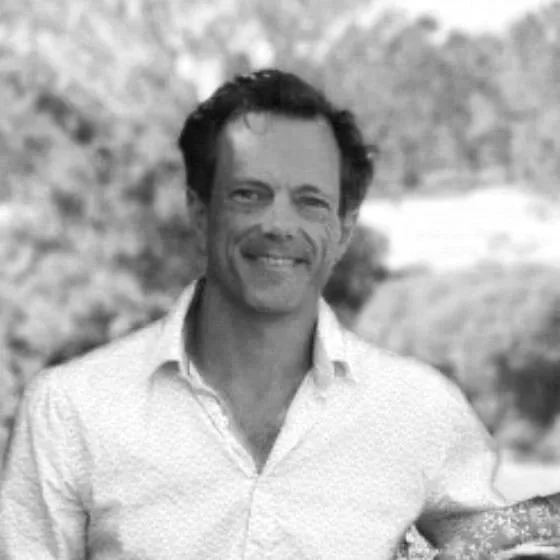 Black and white photo of a smiling man with dark, curly hair outdoors, wearing a patterned button-down shirt.