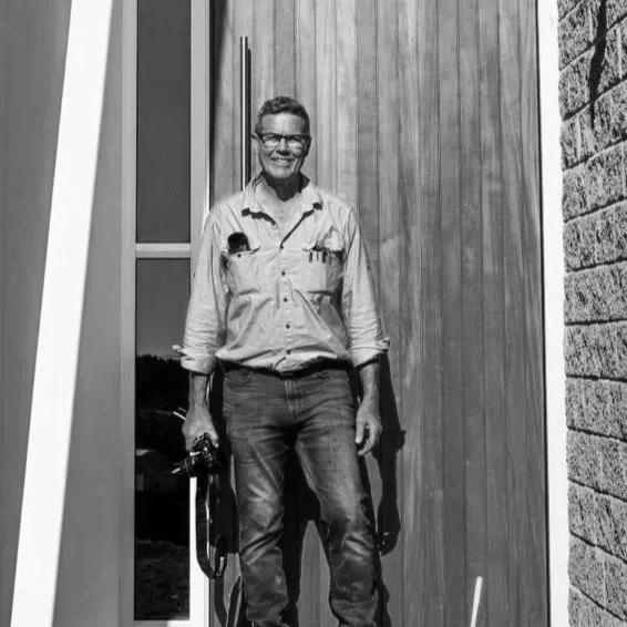 A woman with glasses standing outdoors in front of a wooden wall, holding a camera, smiling.