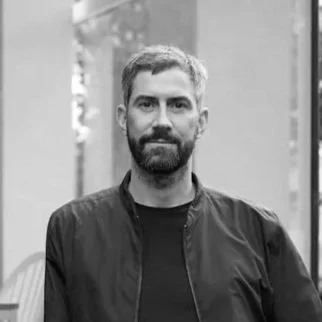 A man with short hair and a beard, wearing a dark jacket, standing outdoors with a blurred background.