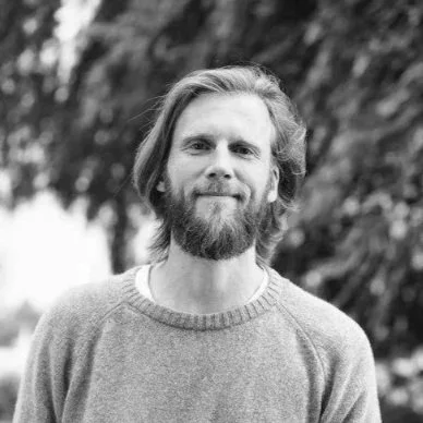 A man with a beard and long hair wearing a gray sweater, standing outdoors with blurred trees in the background.