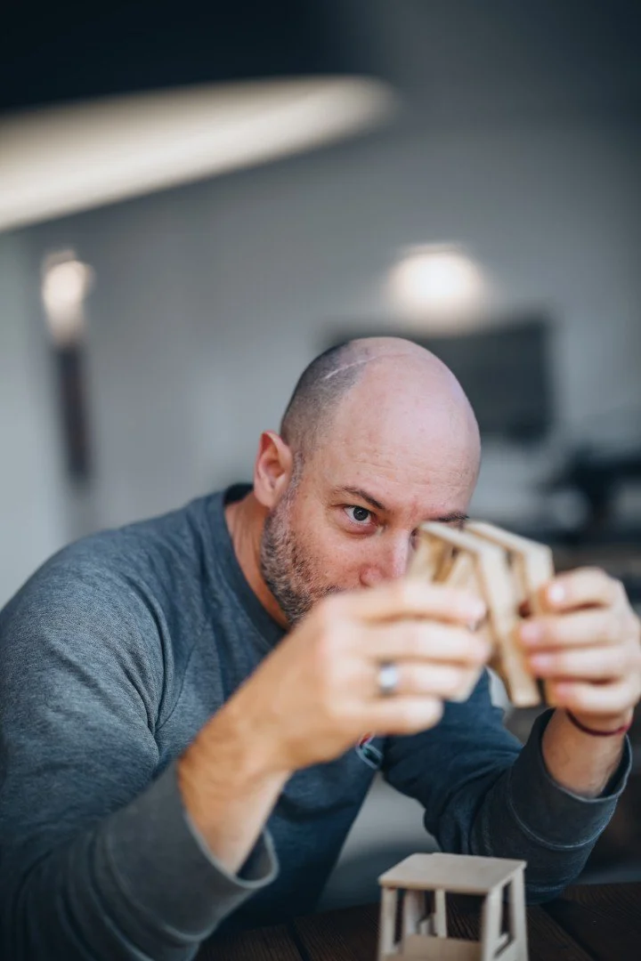 A bald man with a beard wearing a gray shirt, intensely focused on a small wooden object he is holding, with another wooden object on the table in front of him, in an indoor setting.