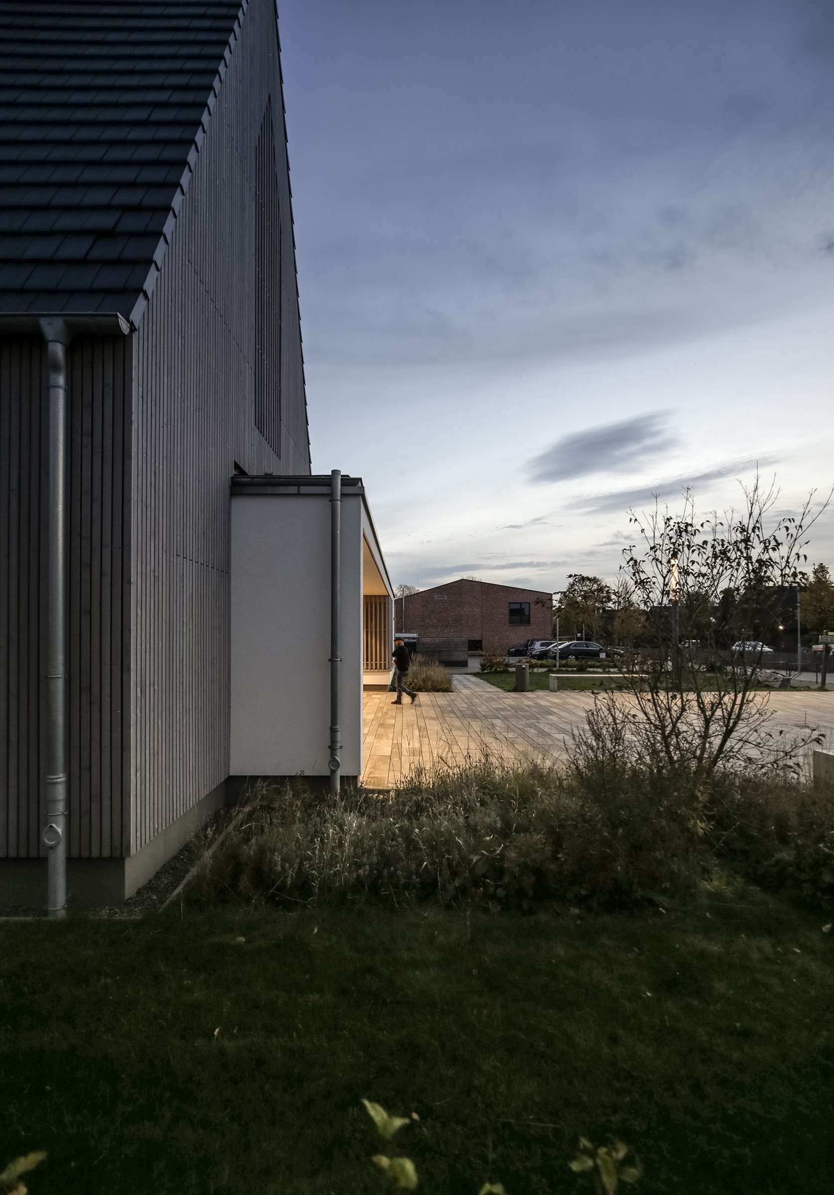 Modern building with dark wooden exterior, outdoor patio with lighting, and a person walking in the distance during early evening or dusk.