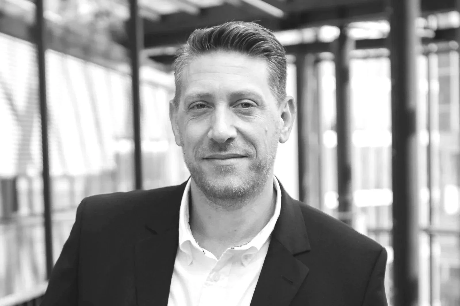 Black and white photo of a man with styled hair, wearing a suit jacket and white shirt, inside a modern glass building with metal framework.
