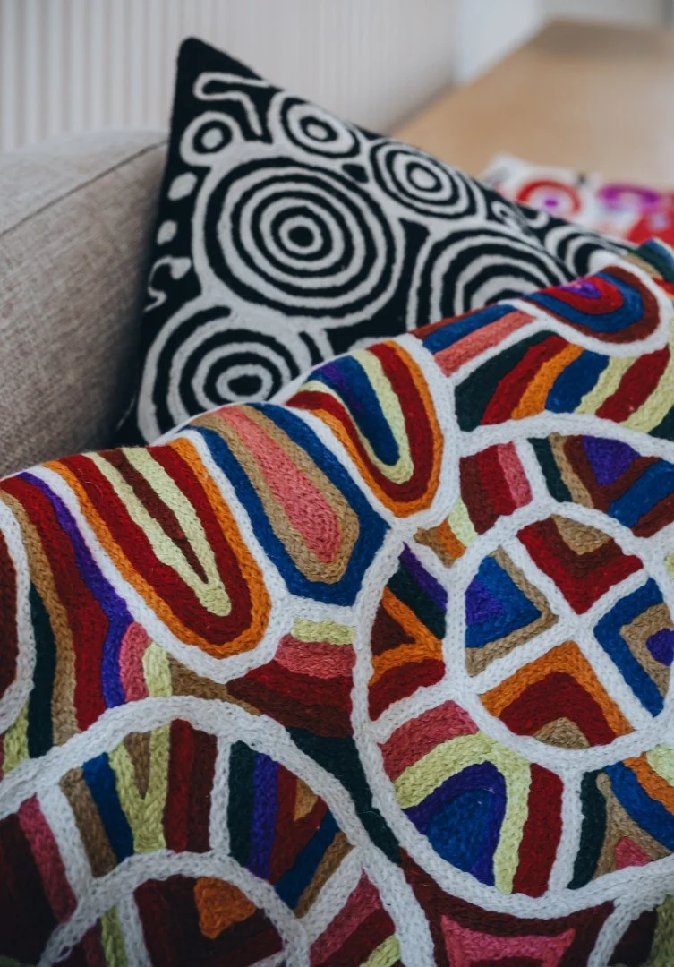 Colorful, abstract-patterned crocheted blanket with concentric circles in red, yellow, orange, purple, and blue in the foreground, and black-and-white patterned pillow with spiral designs resting on a beige couch in the background.