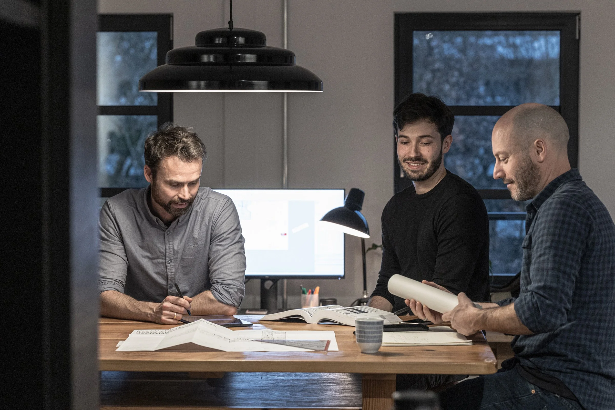 Three men are sitting at a wooden table with documents and books, working together in a modern office with large windows and a computer monitor in the background.