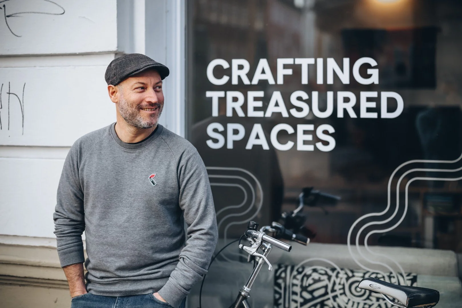 A man with a beard and gray sweater standing outside a shop with a bicycle, smiling, with a window sign that says 'Crafting Treasured Spaces'.