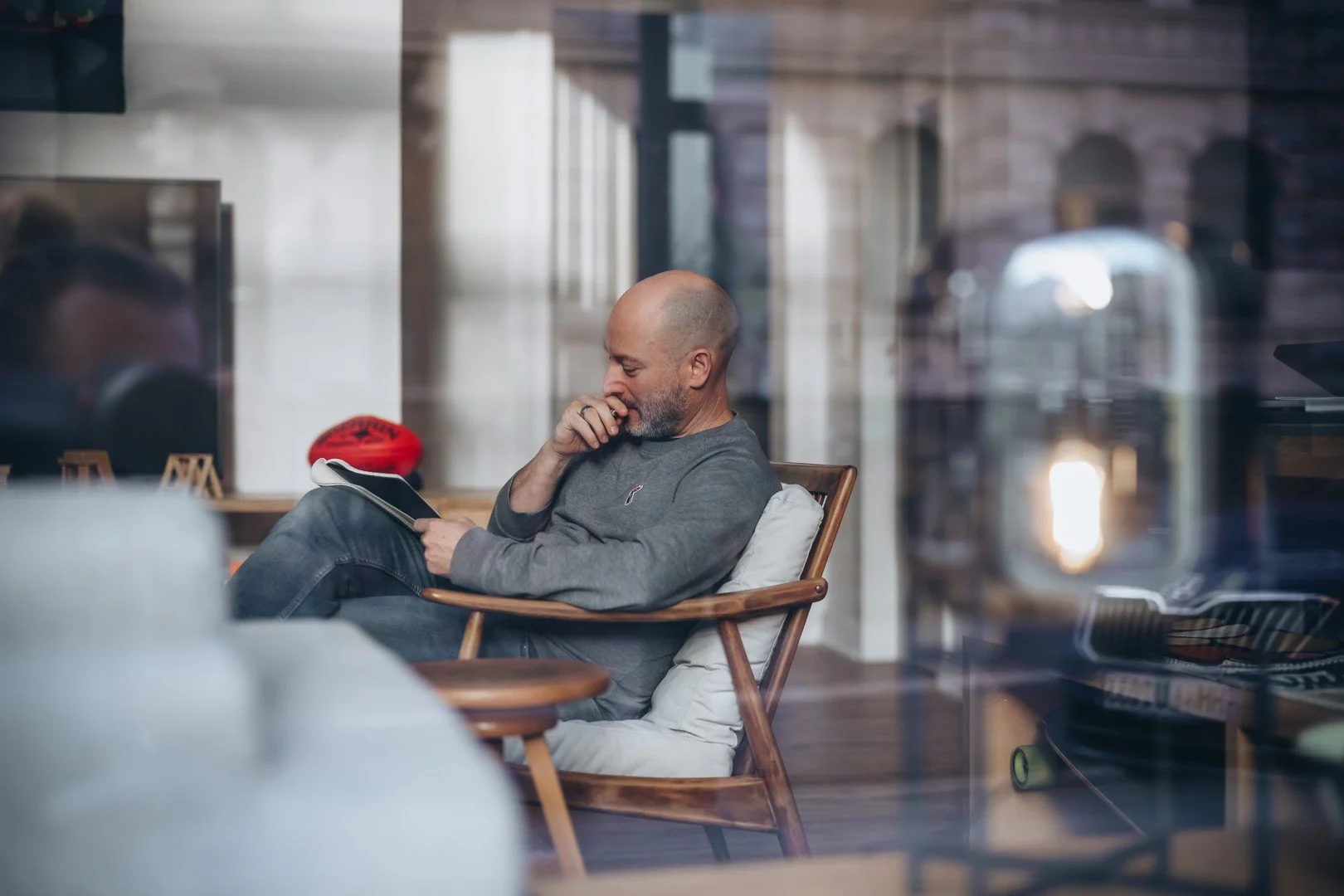 A man with a beard and bald head sits on a wooden chair reading a book inside a cozy room, seen through a window reflective surface.