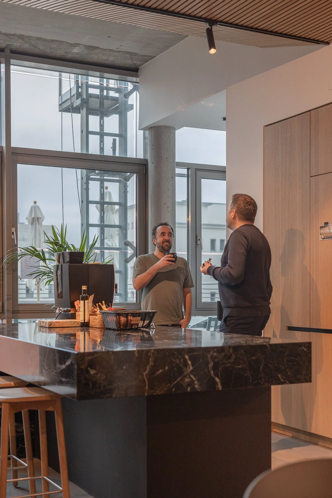 Two men conversing in a modern kitchen with a black marble countertop, a plant, and large windows showing a cityscape and an outdoor elevator structure.