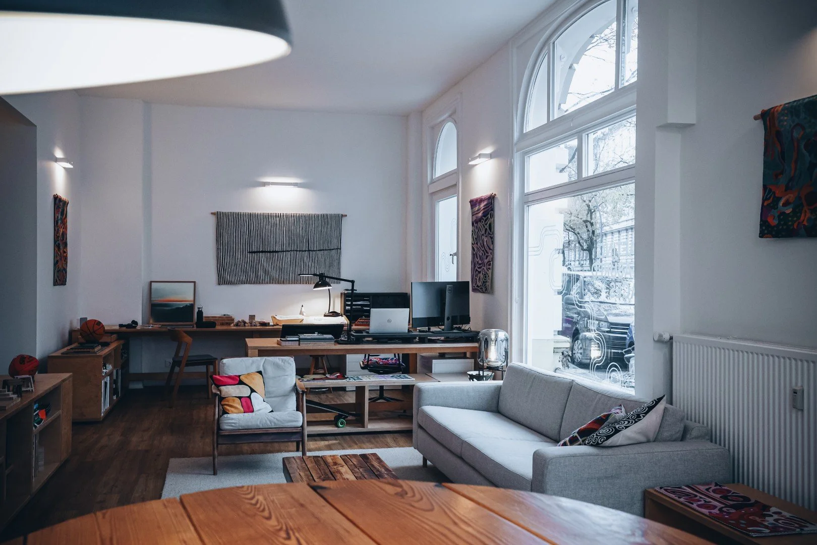 Living room with large windows, white walls, a gray sofa with patterned pillows, a wooden coffee table, a desk with computer monitors, a chair, and abstract wall art.