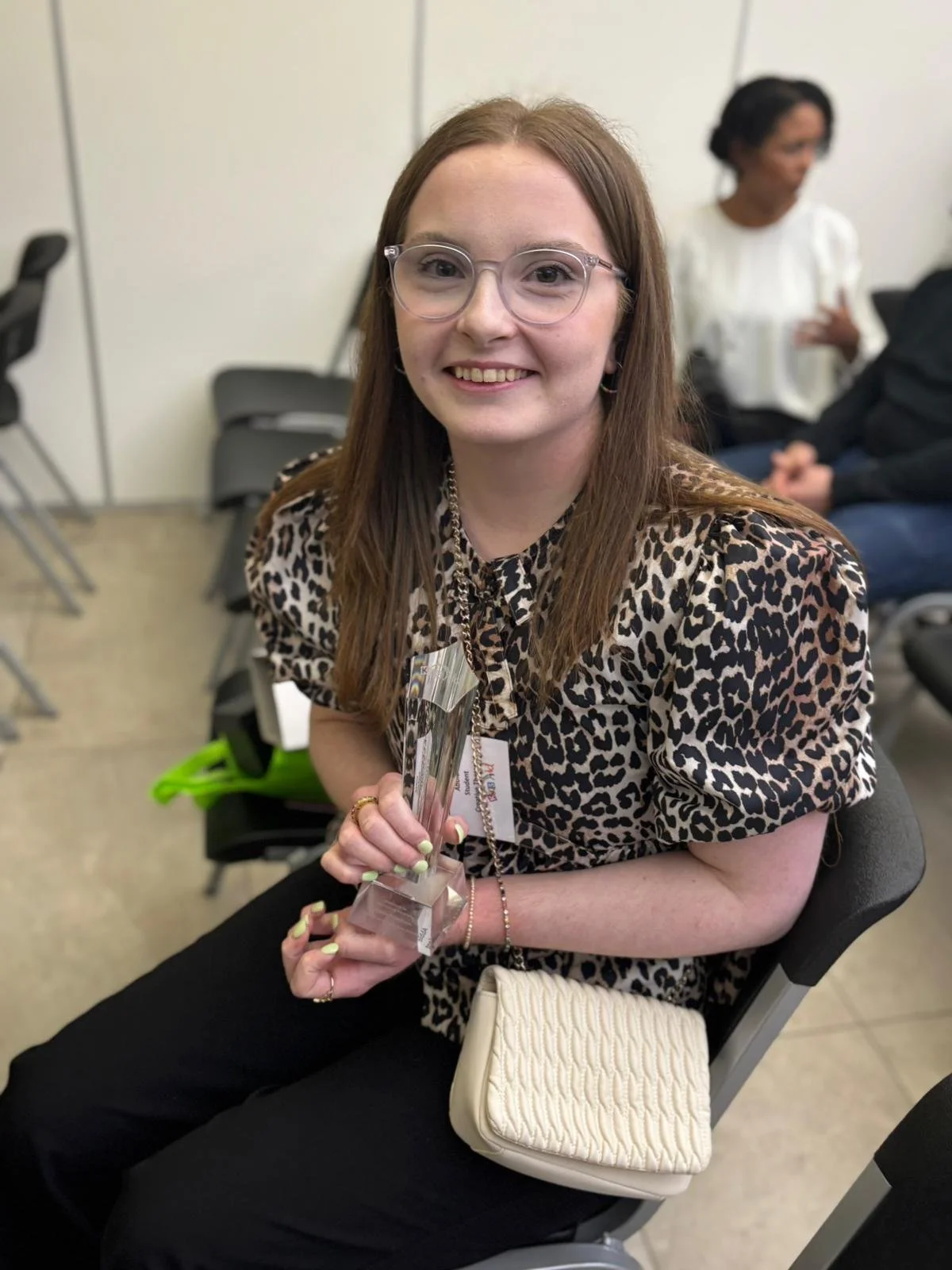 A young woman with glasses and a leopard print top smiling and holding an award, sitting in a chair at an indoor event, with other attendees in the background.