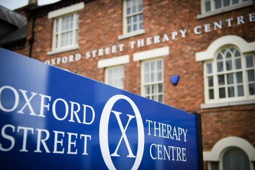 Blue sign for Oxford Street Therapy Centre with white text, in front of a brick building with white window frames.