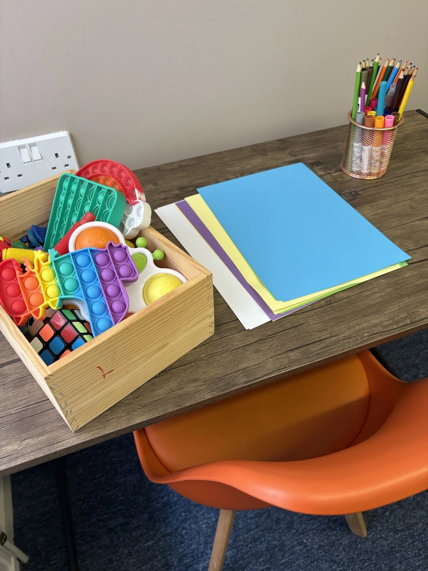 A classroom desk with a wooden container holding colorful fidget toys, a stack of blue, yellow, and purple construction paper, and a clear container filled with multicolored markers. An orange chair is partially visible beneath the desk, and the wall behind it is plain.