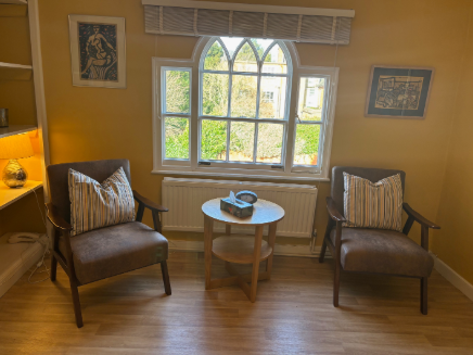 Living room with two brown armchairs with striped cushions facing a window, a small round wooden table with a decorative bowl in the center, and framed artwork on the yellow walls.
