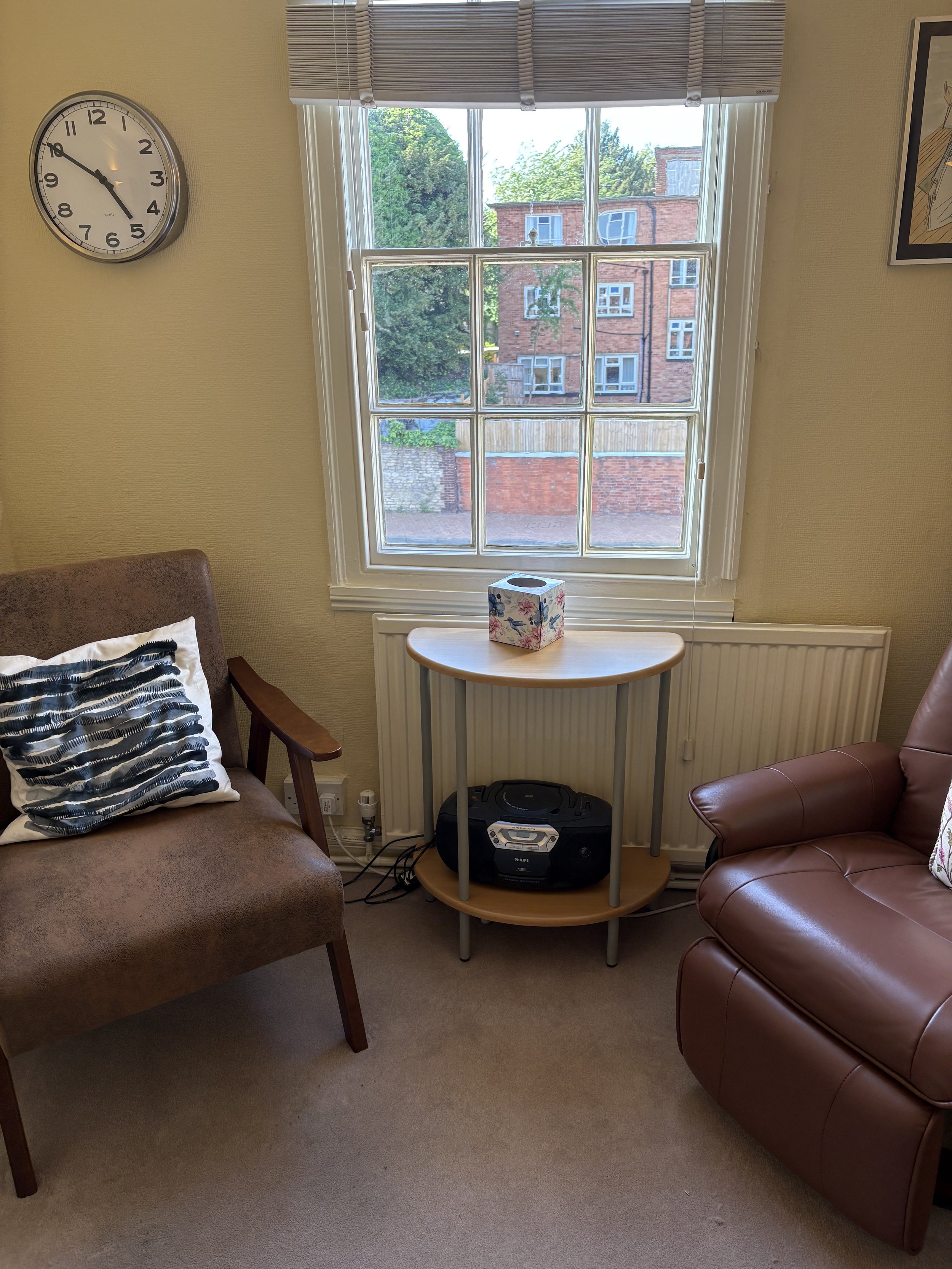 A cozy living room corner with a window overlooking a brick building and trees outside. There are two armchairs, one brown with a patterned pillow and the other in dark red leather. A small wooden table with a floral tissue box sits between the chairs, and a stereo system is underneath the table. A clock on the wall shows the time as approximately 9:48.