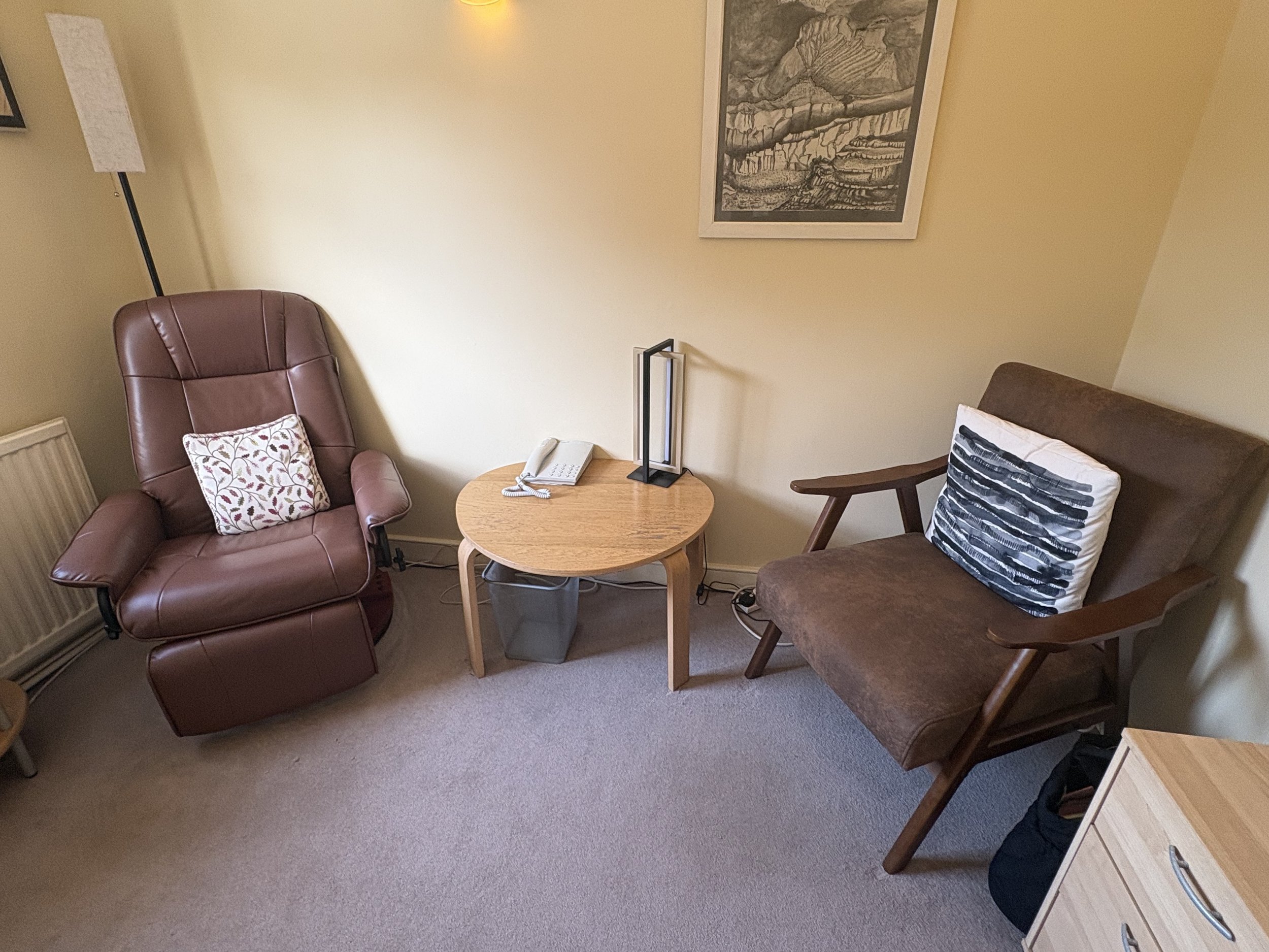 A cozy sitting area with a brown leather armchair, a wooden side table with a white telephone and a modern black desk lamp, another wooden chair with a black and white patterned pillow, a framed black-and-white landscape picture on the yellow wall, and a floor lamp with a white shade.