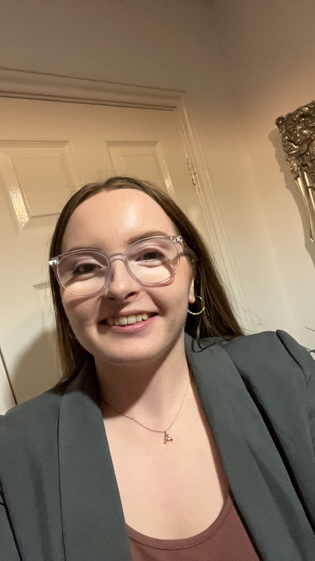 A woman with brown hair, wearing glasses, earrings, a gray blazer, a brown top, and a necklace, smiling for a selfie indoors.