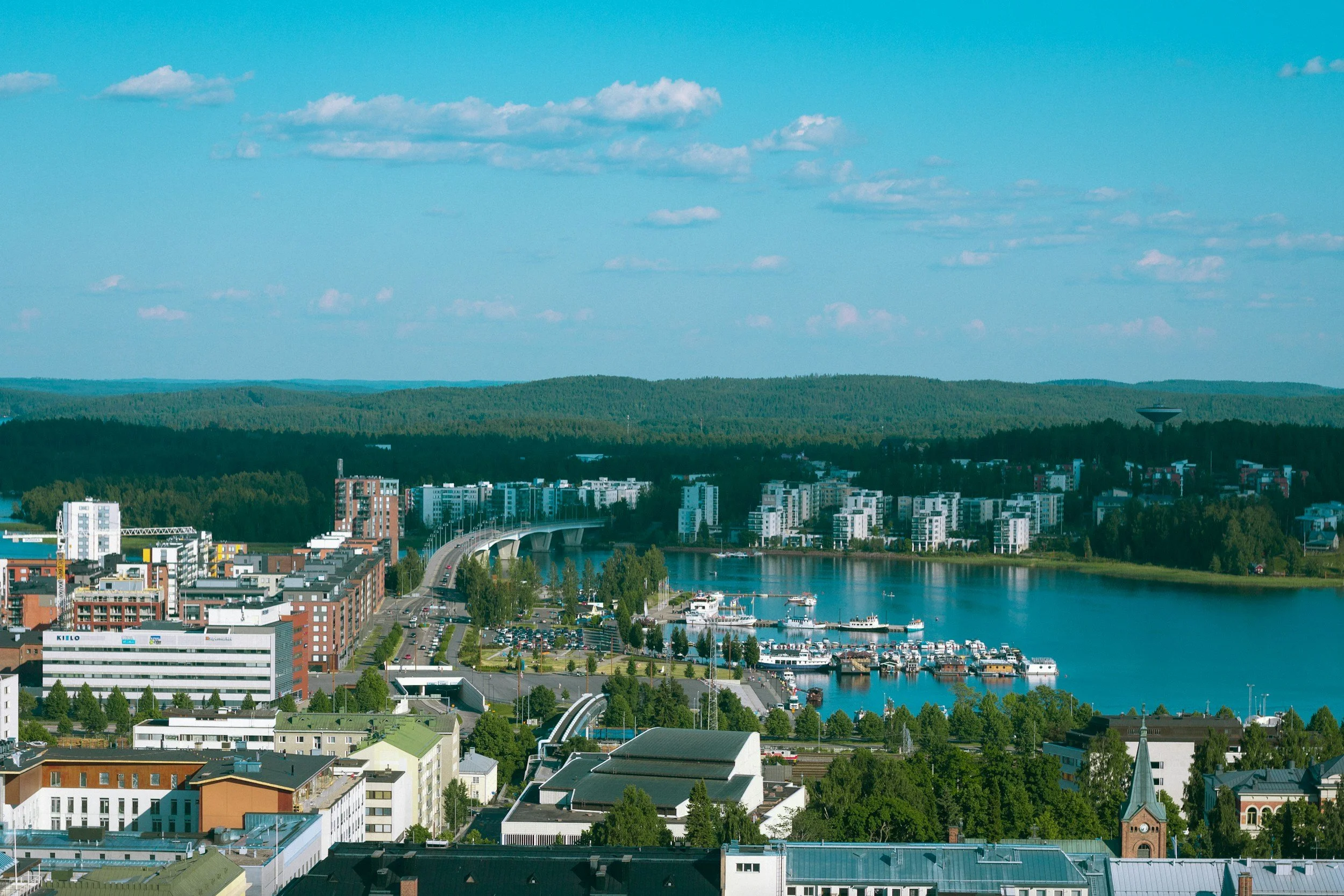 Kuva kaupunkinäkymä järven rannalta, useita rakennuksia, veneitä ja silta, taustalla metsää ja sininen taivas pilvinä.