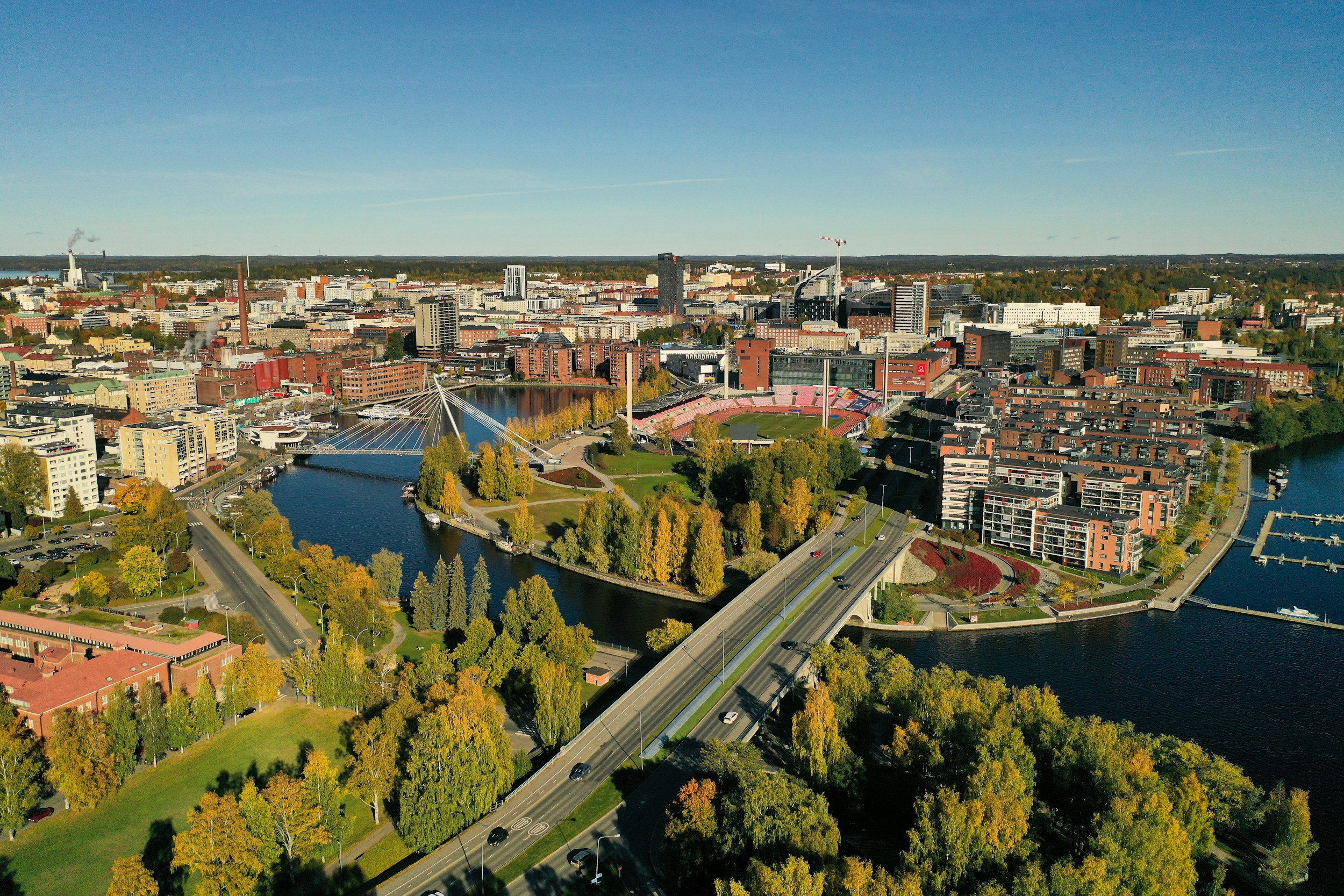 Kattava ilmakuva kaupungin keskustasta, jossa on joki, siltoja, puistoja ja korkeita rakennuksia, selkeällä säällä.