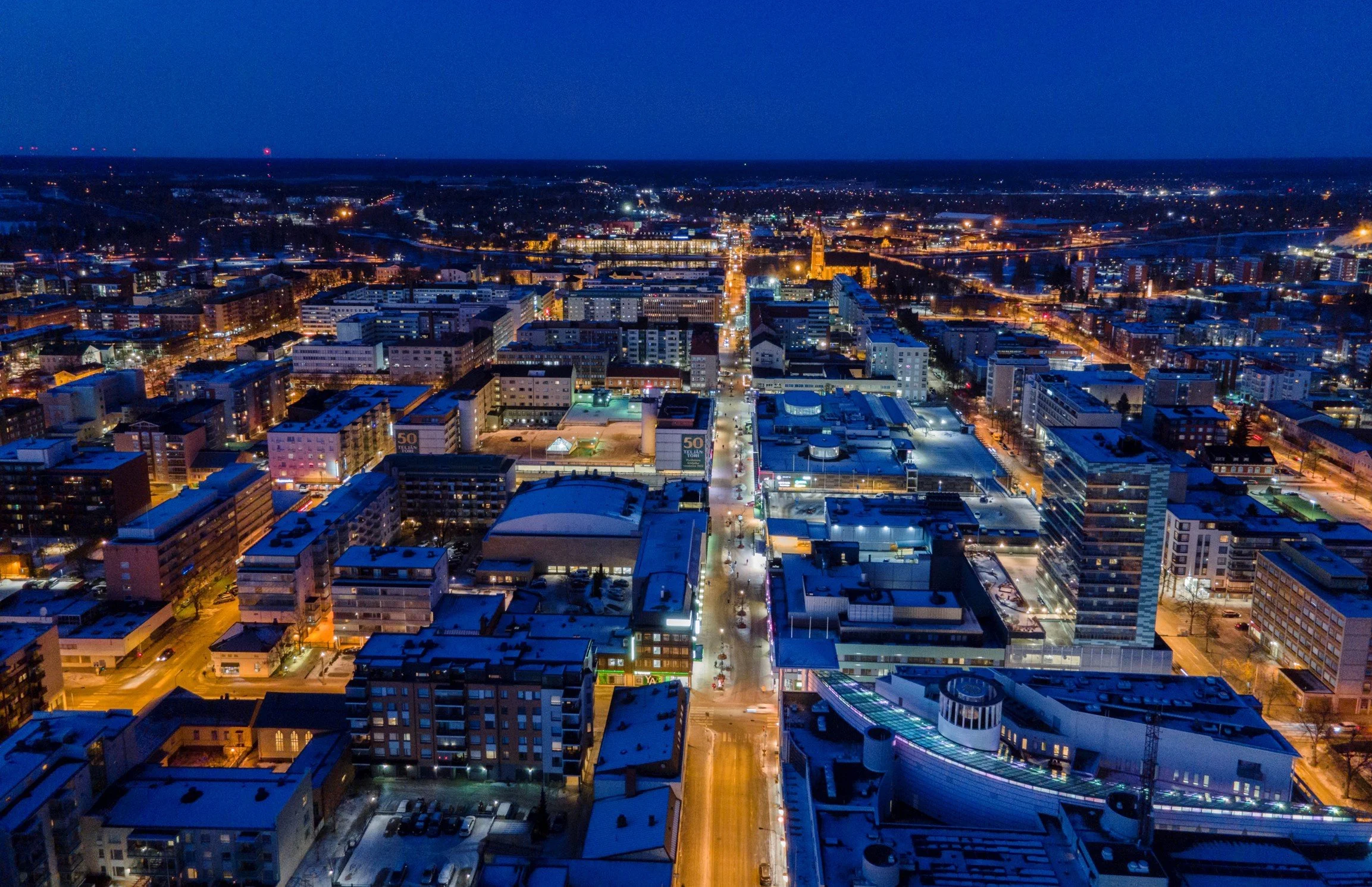 Kaupunkinäkymä iltavalossa, korkeita rakennuksia ja katuvaloja, lumipeite katolla ja teillä, taivas pimeä.