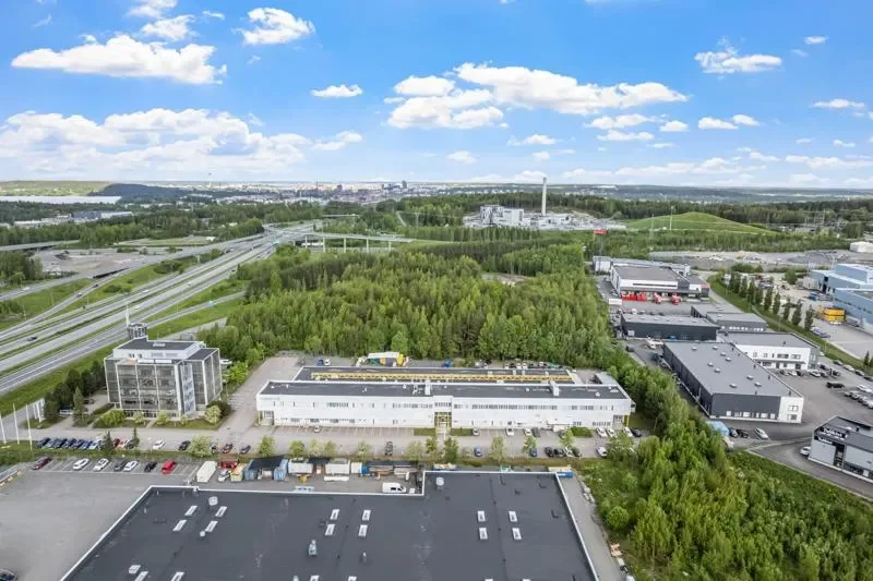 Kattovalokuva teollisuusalueesta, runsaasti rakennuksia, parkkipaikkoja ja metsää, taivaalla pilviä.