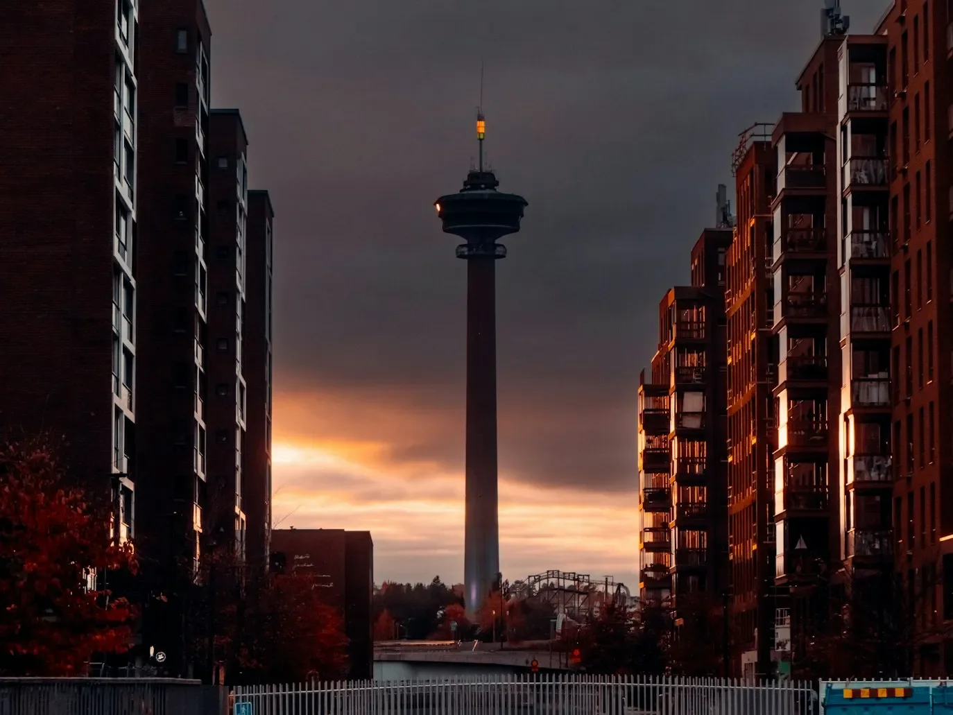 Kuva kaupunkimaisemasta auringonlaskun aikaan, sisältää korkean televisio- tai radiotorni keskellä, ympärillä kerrostaloja ja rakennuksia.