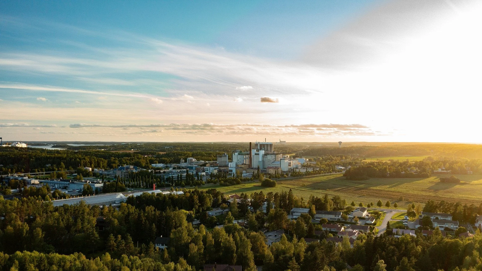 Valokuva kaupungista ja tehtaasta metsäisen alueen keskellä auringon laskiessa.