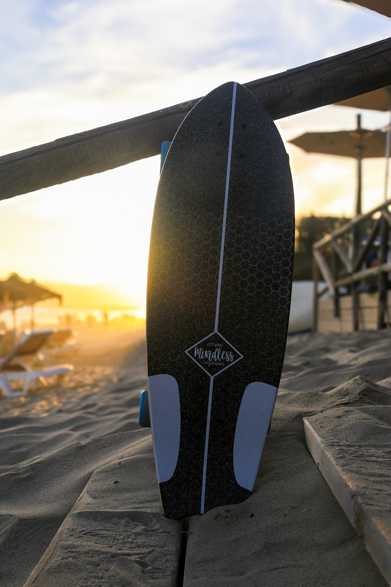 A black and blue wakeboard with the brand name 'Mindless' standing upright on a sandy beach near a wooden structure, during sunset with a lounge chair and umbrella in the background.