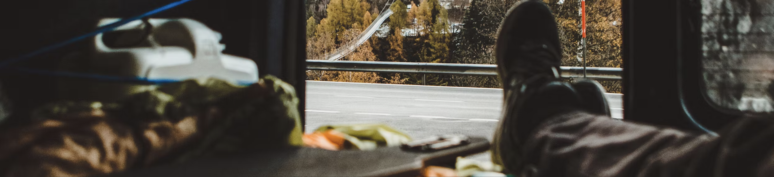 View from inside a vehicle showing a person's legs and feet outside on a road with trees in the background.