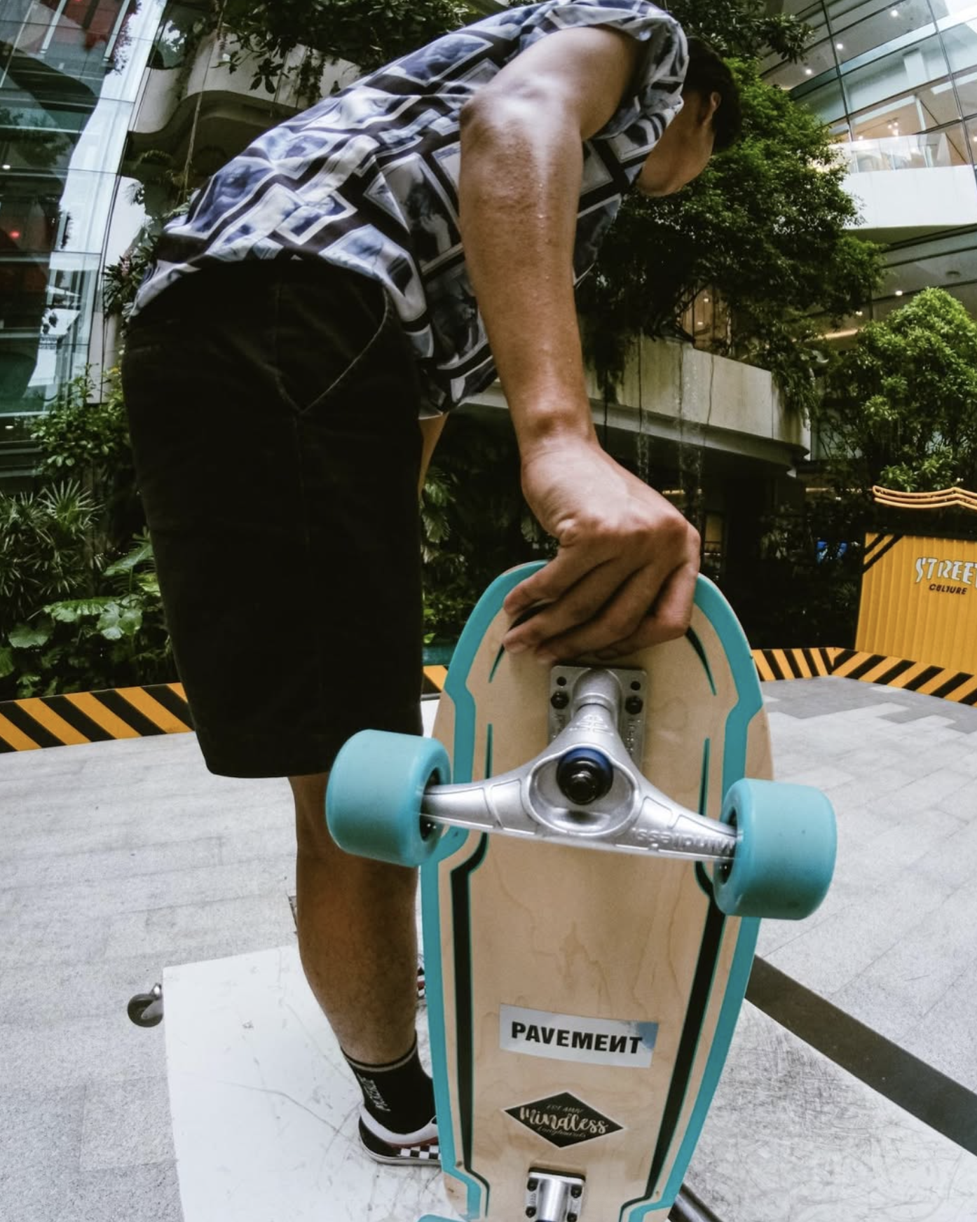 Person adjusting skateboard outdoors in urban setting with modern glass building and greenery in background.
