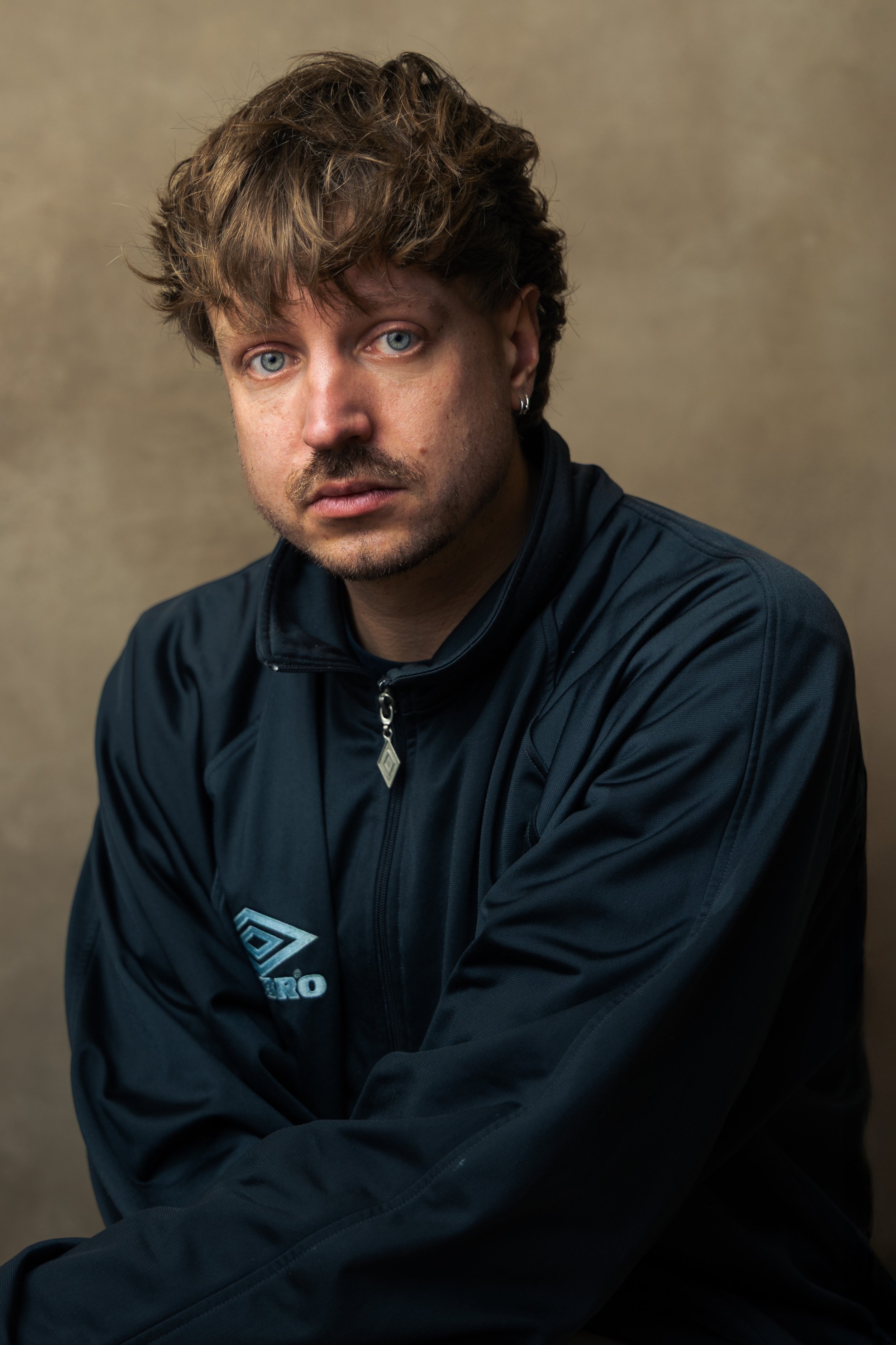 A young man with light brown, slightly curly hair and blue eyes, wearing a dark blue sports jacket with a zippered collar and a logo on the chest, sitting against a neutral beige background.