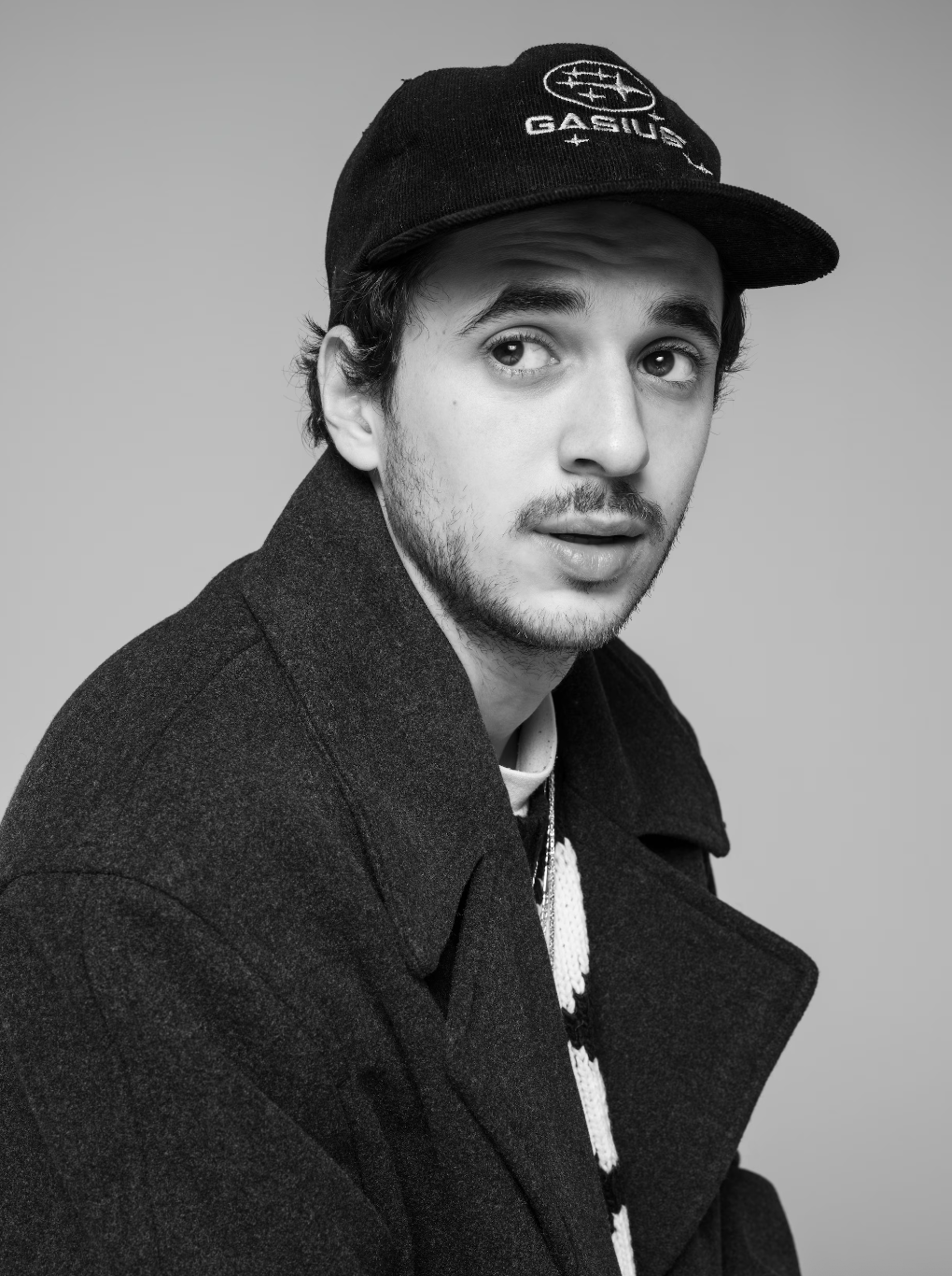 A young man with light skin, dark hair, and a beard, wearing a dark coat and a black cap with a logo, looking to the side, in black and white.