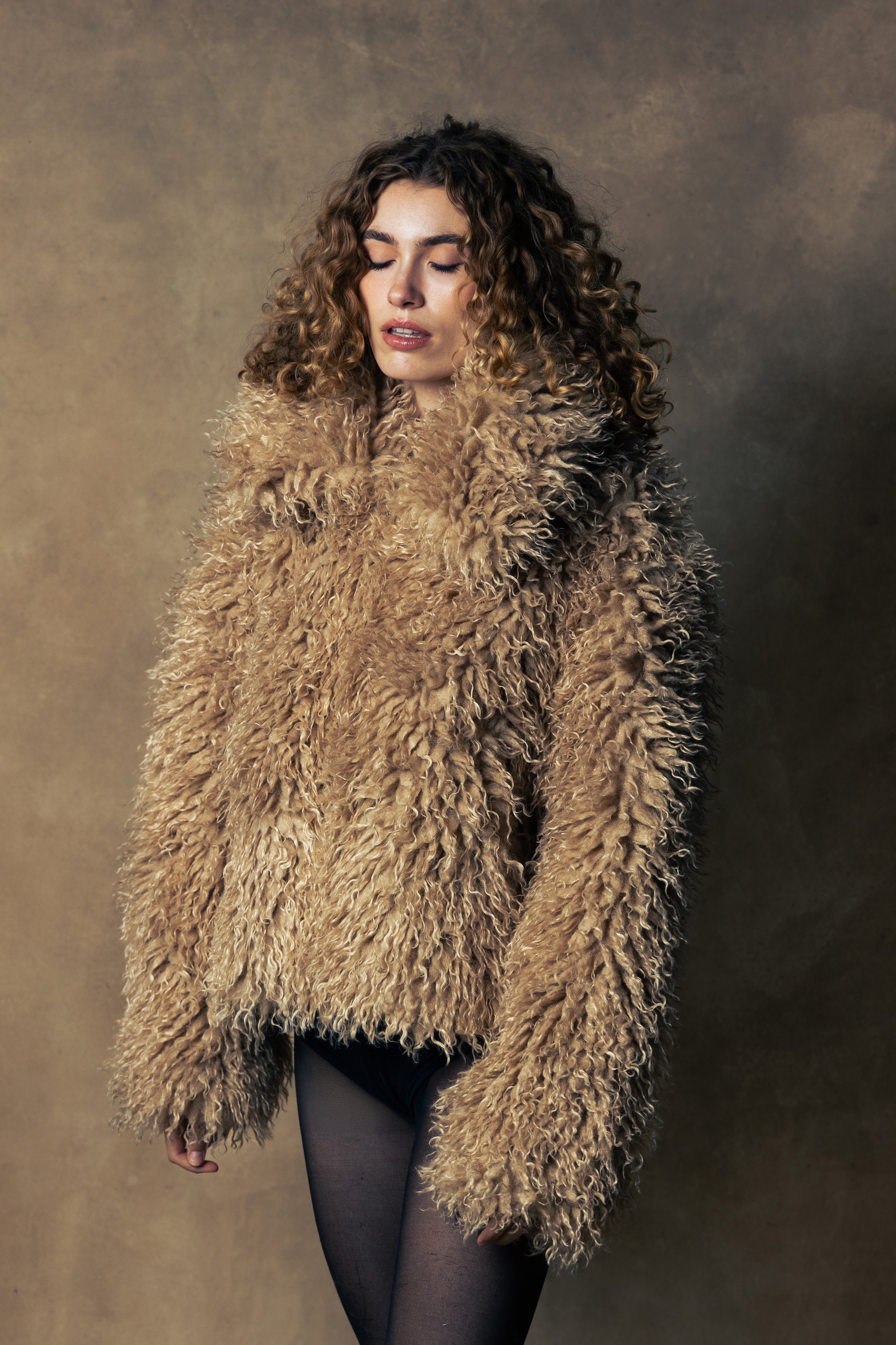 A woman with curly hair wearing a fluffy brown coat and black tights standing against a plain background with her eyes closed.