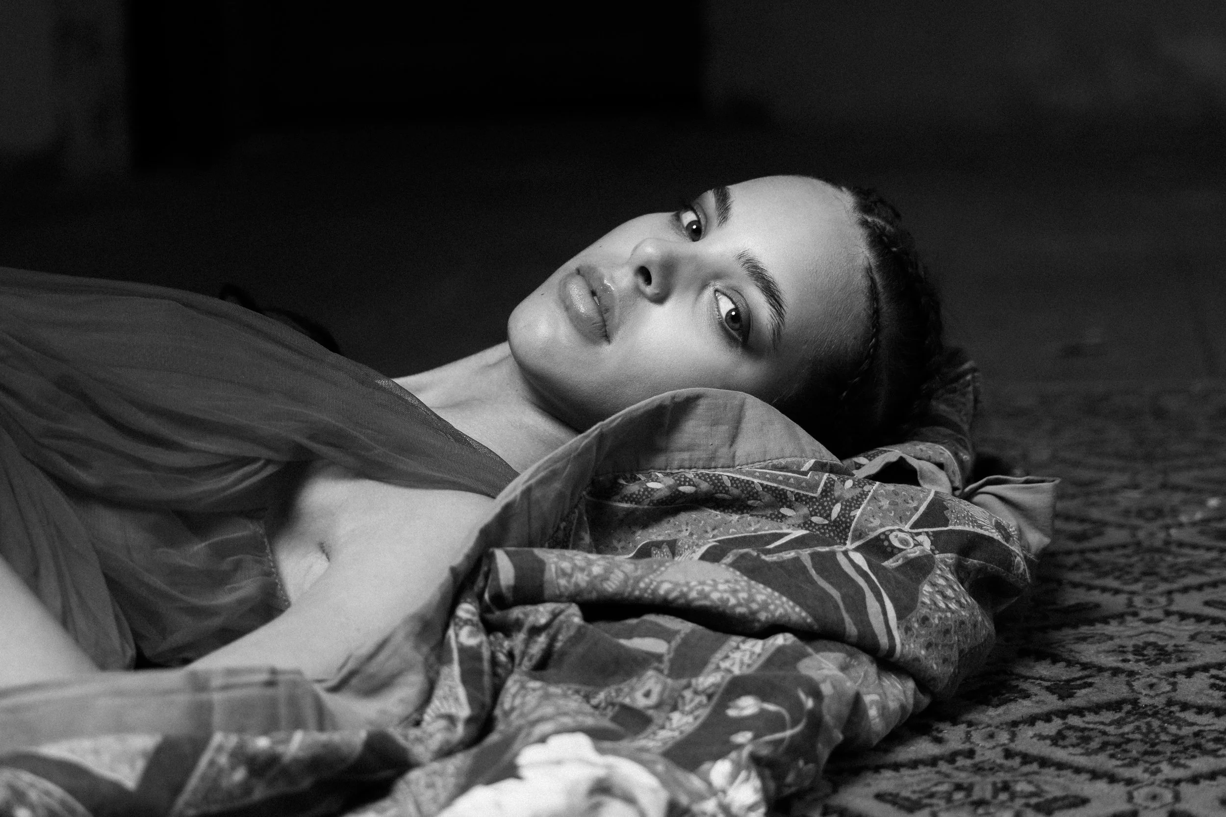 A woman with braided hair lying on a patterned blanket, looking at the camera in black and white.