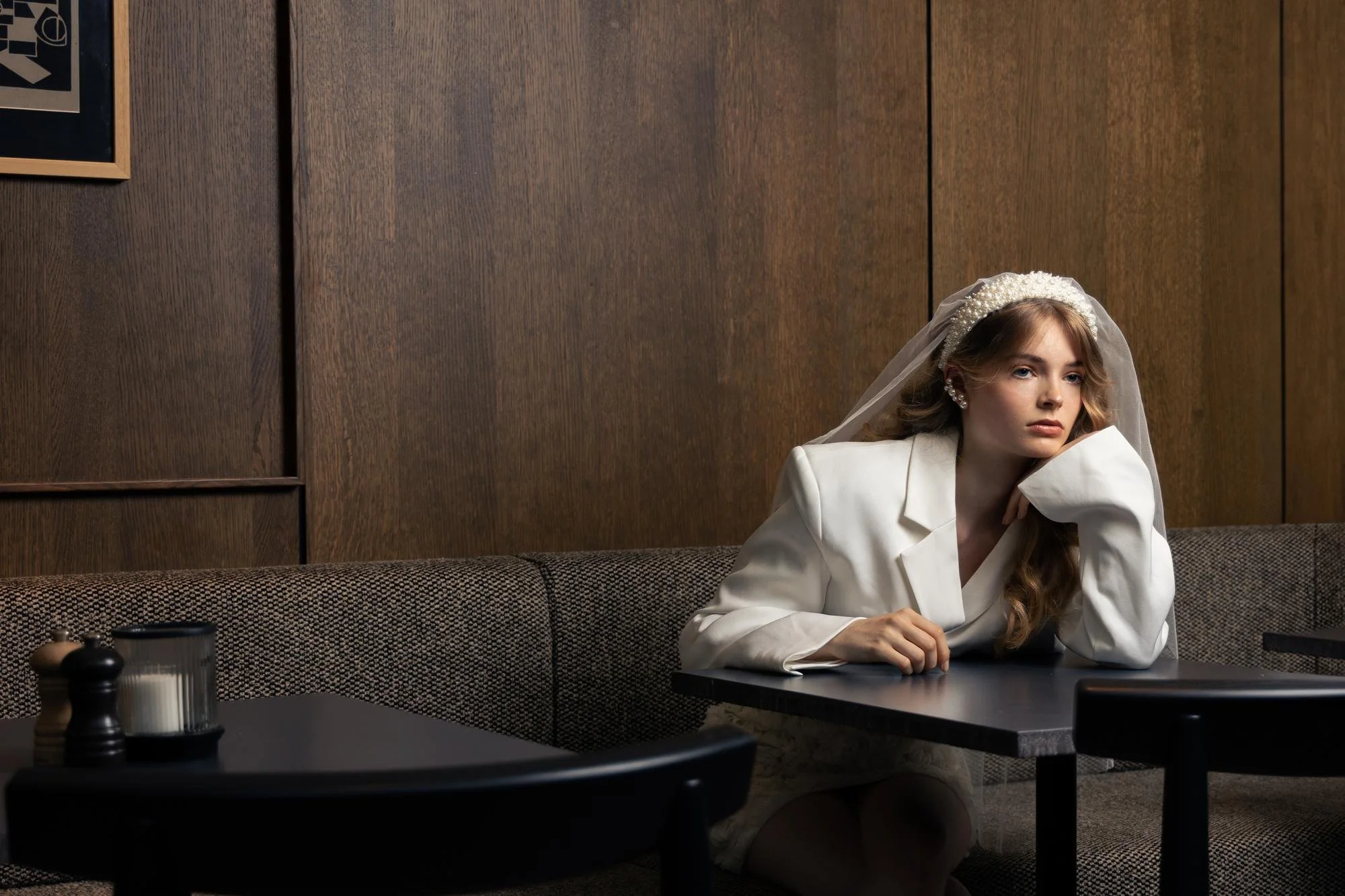 A young woman with long wavy hair, wearing a white blazer, with a pearl headband, veil, and earrings, sitting alone at a table in a restaurant or cafe, resting her head on her hand, looking contemplative.