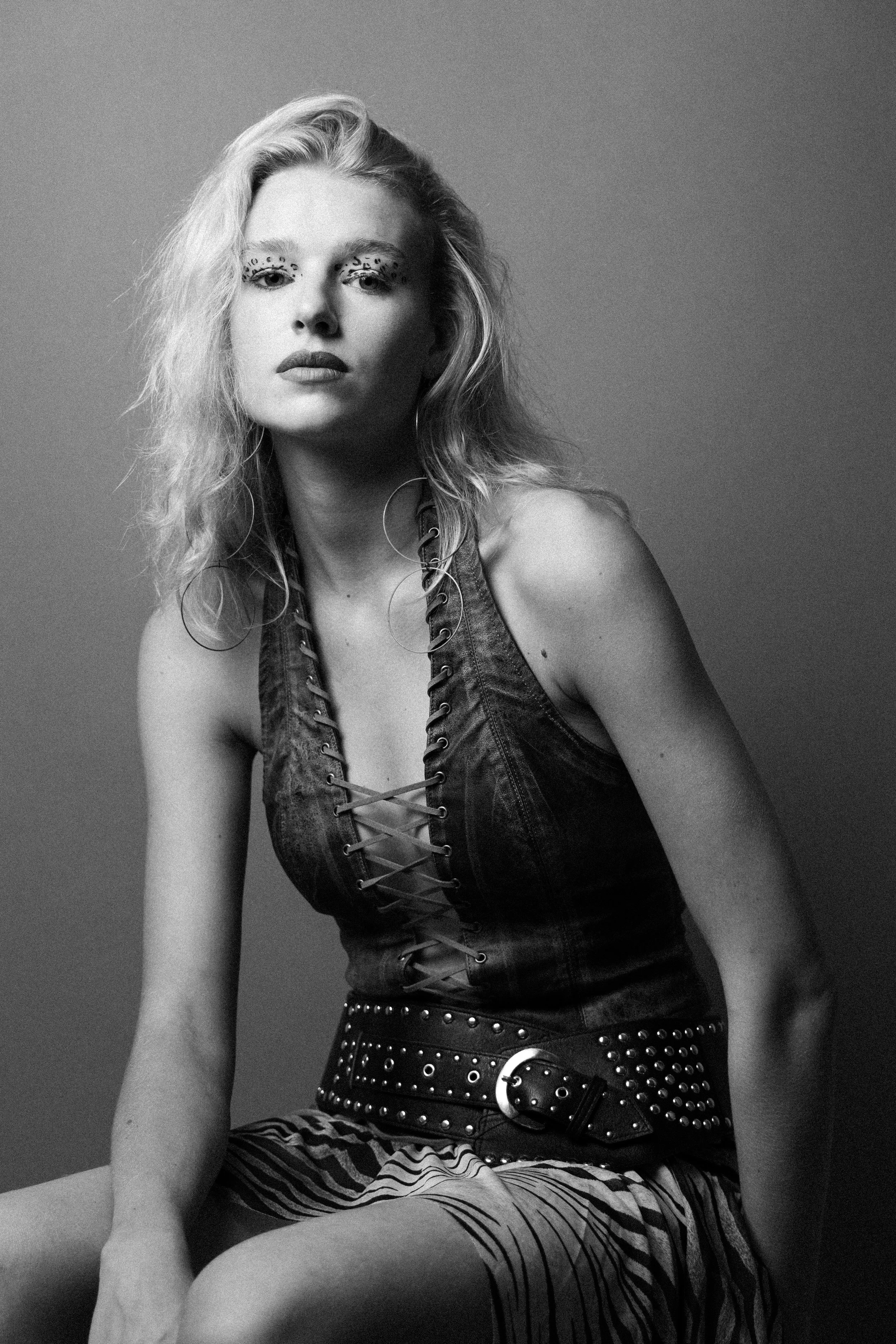 Black and white portrait of a woman with wavy blonde hair wearing a sleeveless leather top with lace-up front, a wide studded belt, and striped shorts, sitting against a plain background.